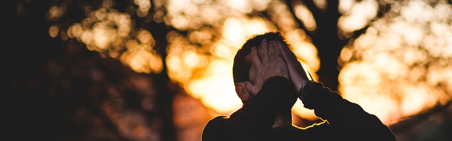 a man holds both hands over his face with trees backlit by the sun going down | risking connection at work | Tempo Therapy and Consulting