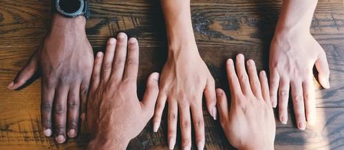 five hands of different colours sit palms down on a wooden table | why self care is not enough in healthcare | Tempo Therapy and Consulting