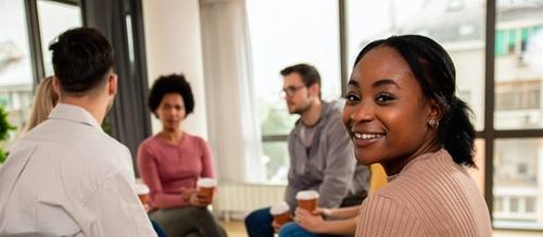 a group of adults sit in discussion as a young woman with brown skin turns to smile at the camera | collective care | Tempo Therapy and Consulting