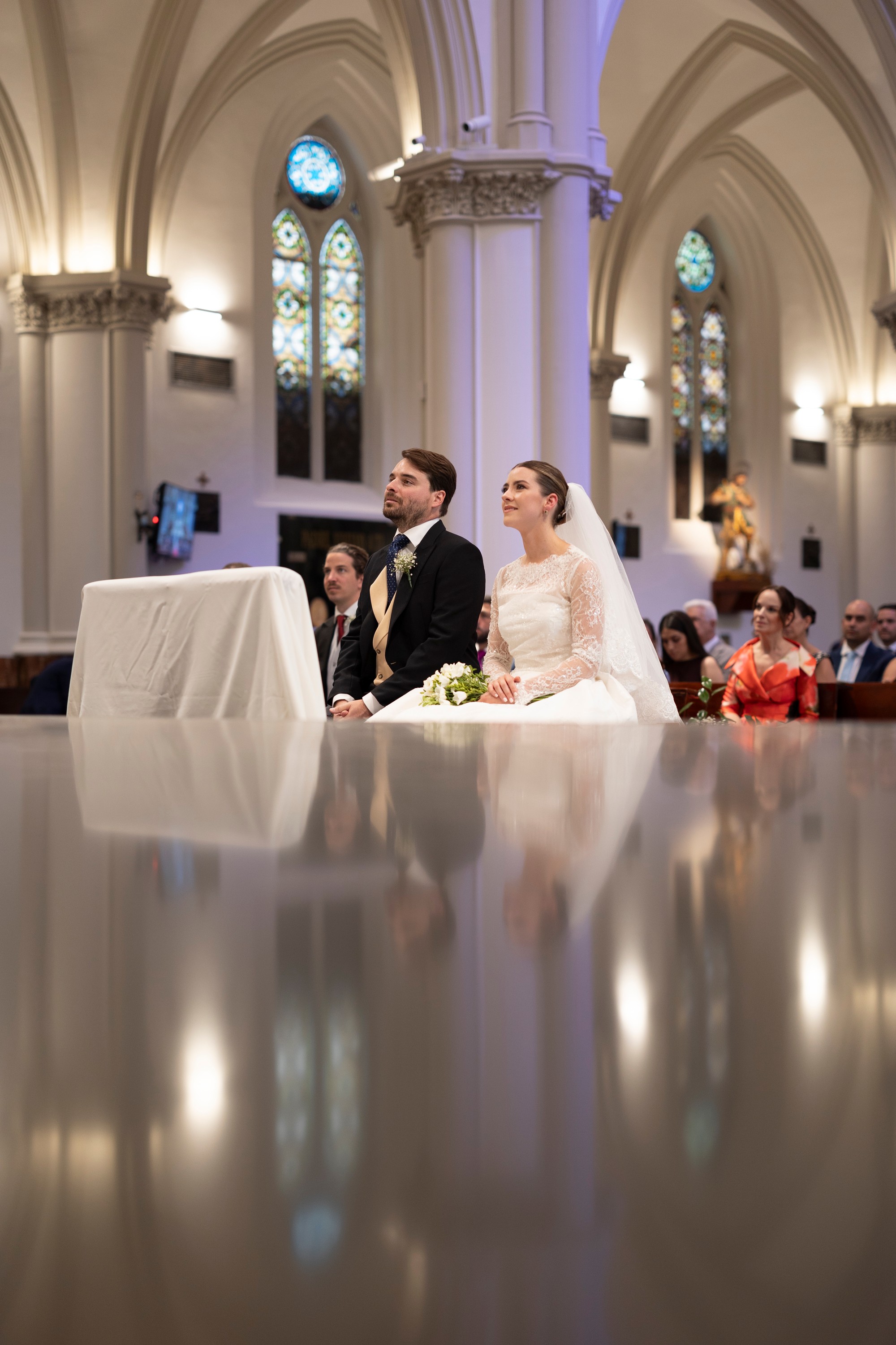 Los novios sentados durante la ceremonia se reflejan en el suelo pulido de la iglesia
