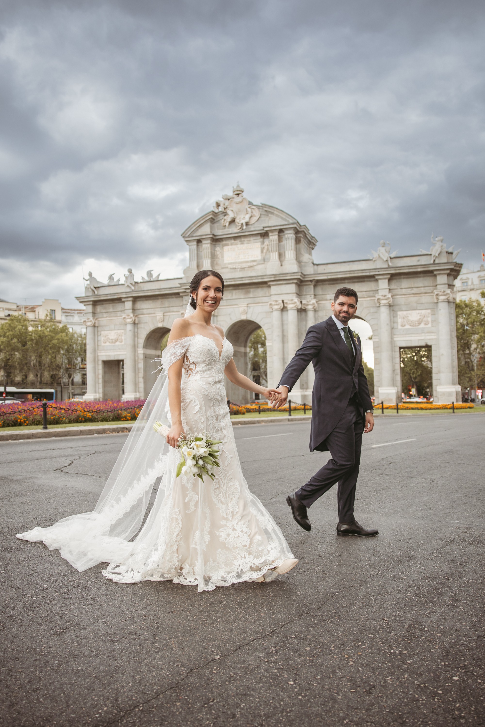 Los novios cruzan la calle de la mano con la Puerta de Alcalá y un cielo de tormenta detrás