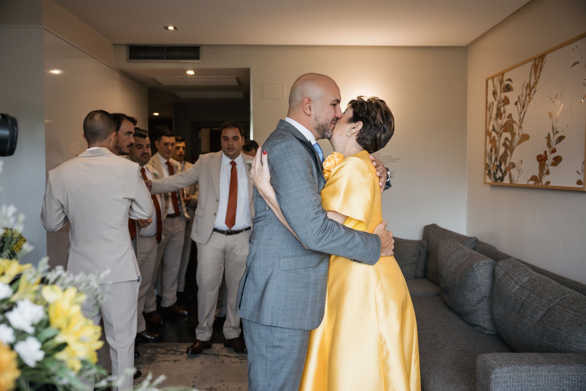 El novio abraza y besa a una invitada de vestido amarillo mientras sus amigos esperan en el pasillo