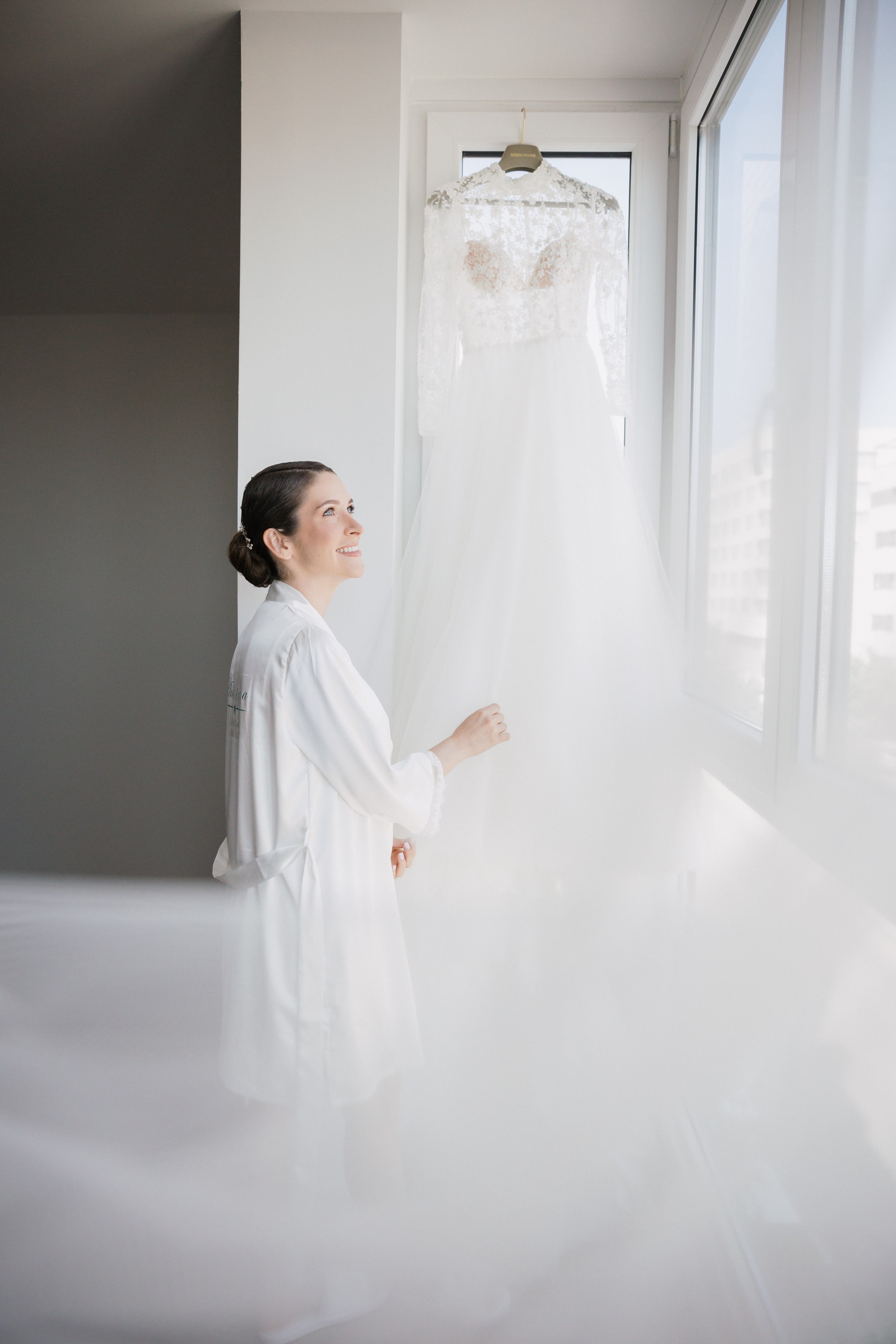 La novia, en bata, mira su vestido colgado junto a la ventana con el velo extendido por el suelo