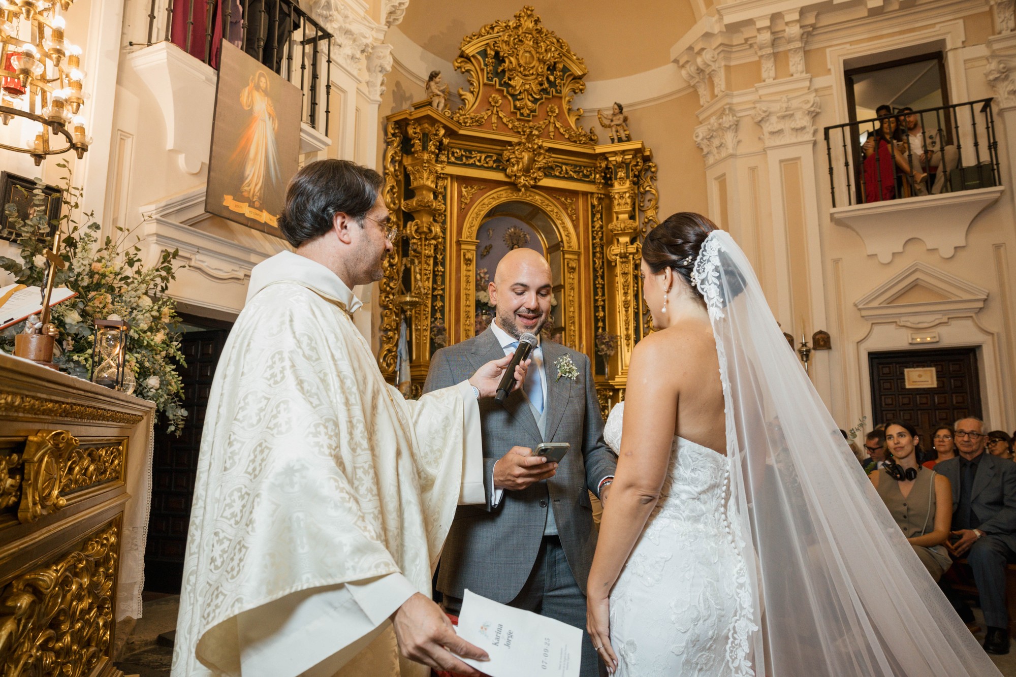 El novio lee sus votos desde el móvil ante el micrófono que le sostiene el sacerdote