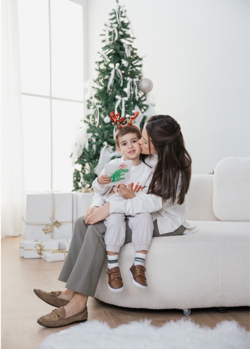 Mother kissing her son, who wears reindeer antlers, on a white sofa in front of the Christmas tree