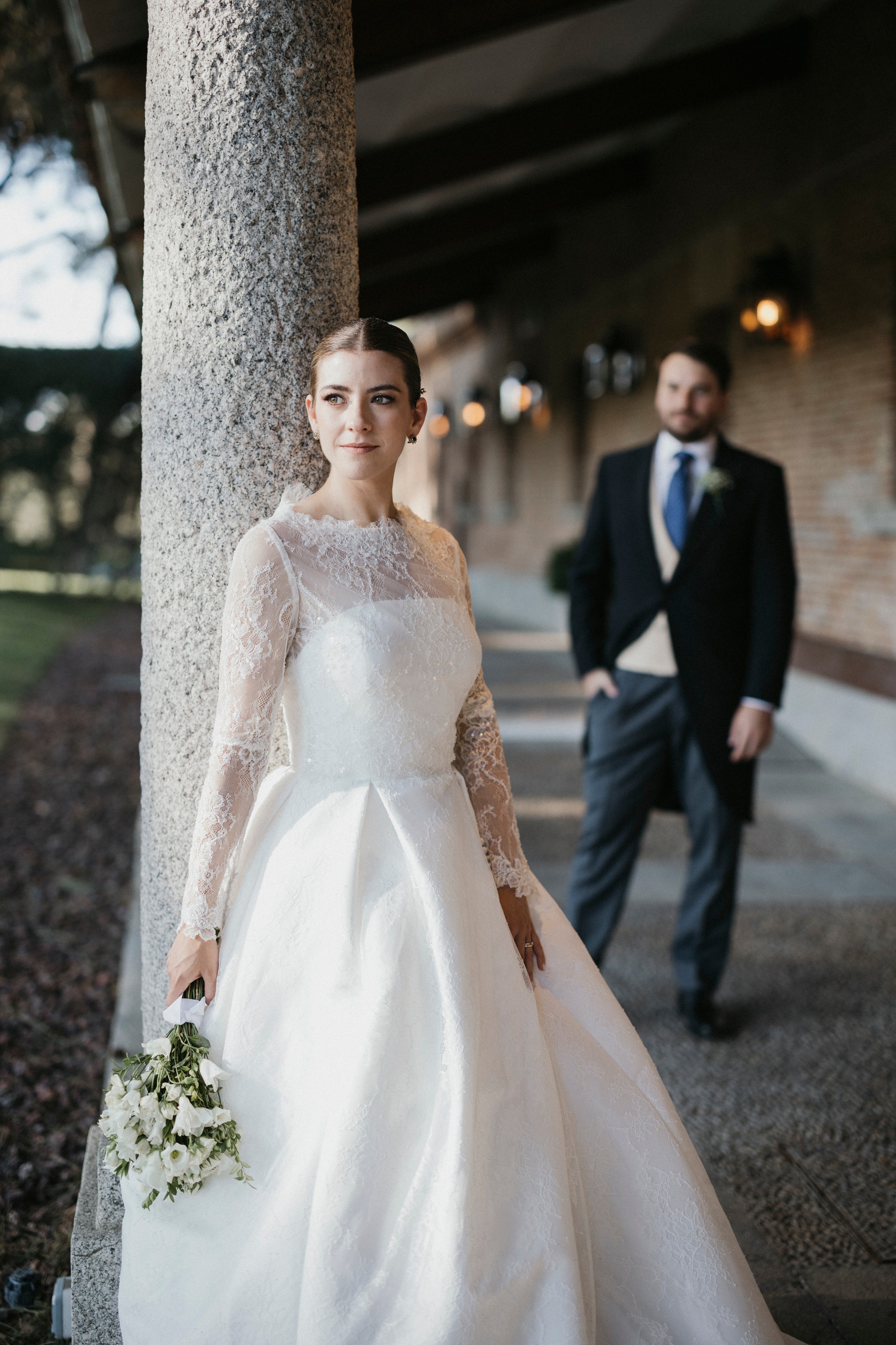 La novia mira a cámara junto a la columna mientras el novio se acerca al fondo