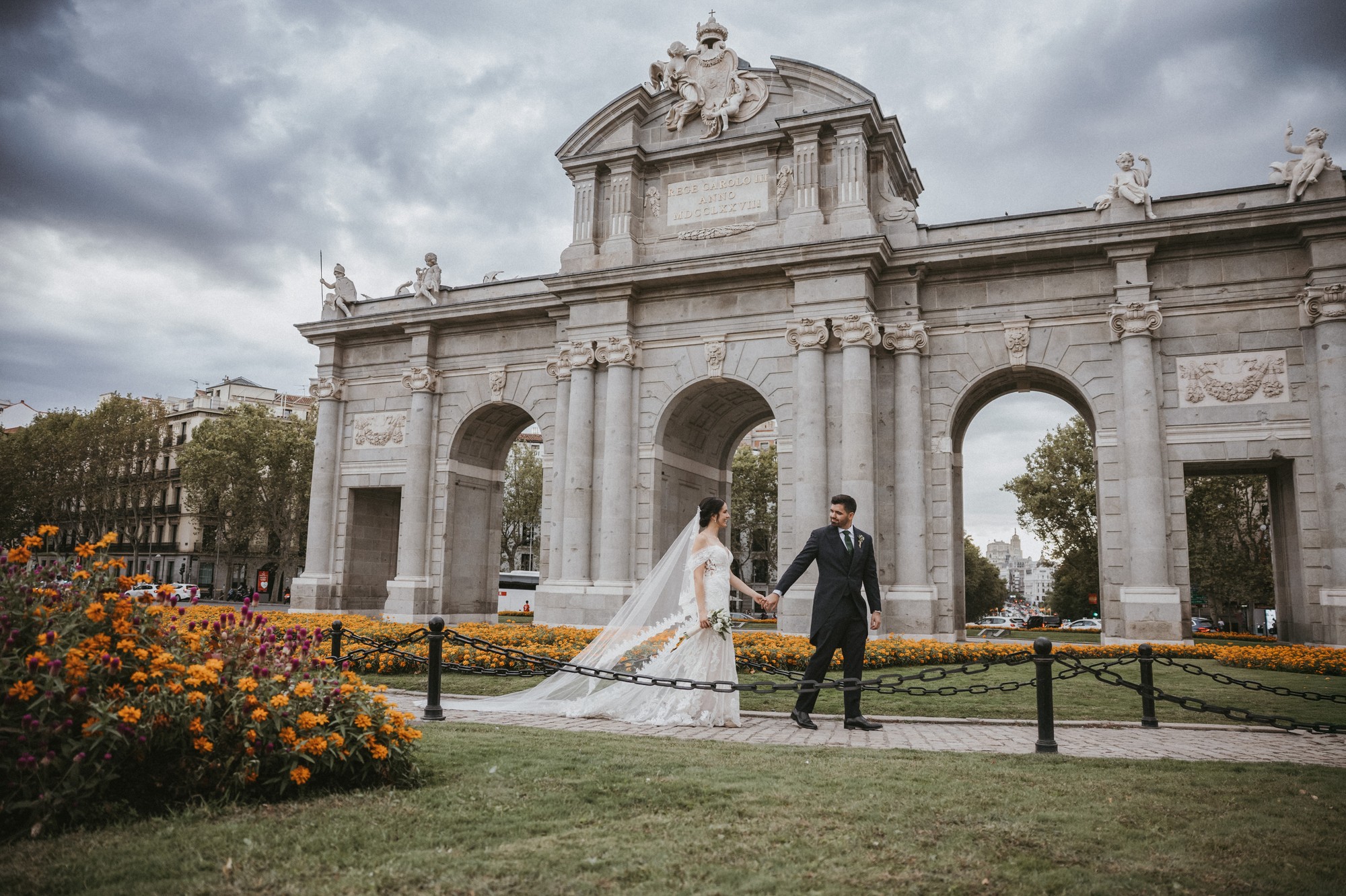 Los novios pasean de la mano ante la Puerta de Alcalá entre los parterres de flores