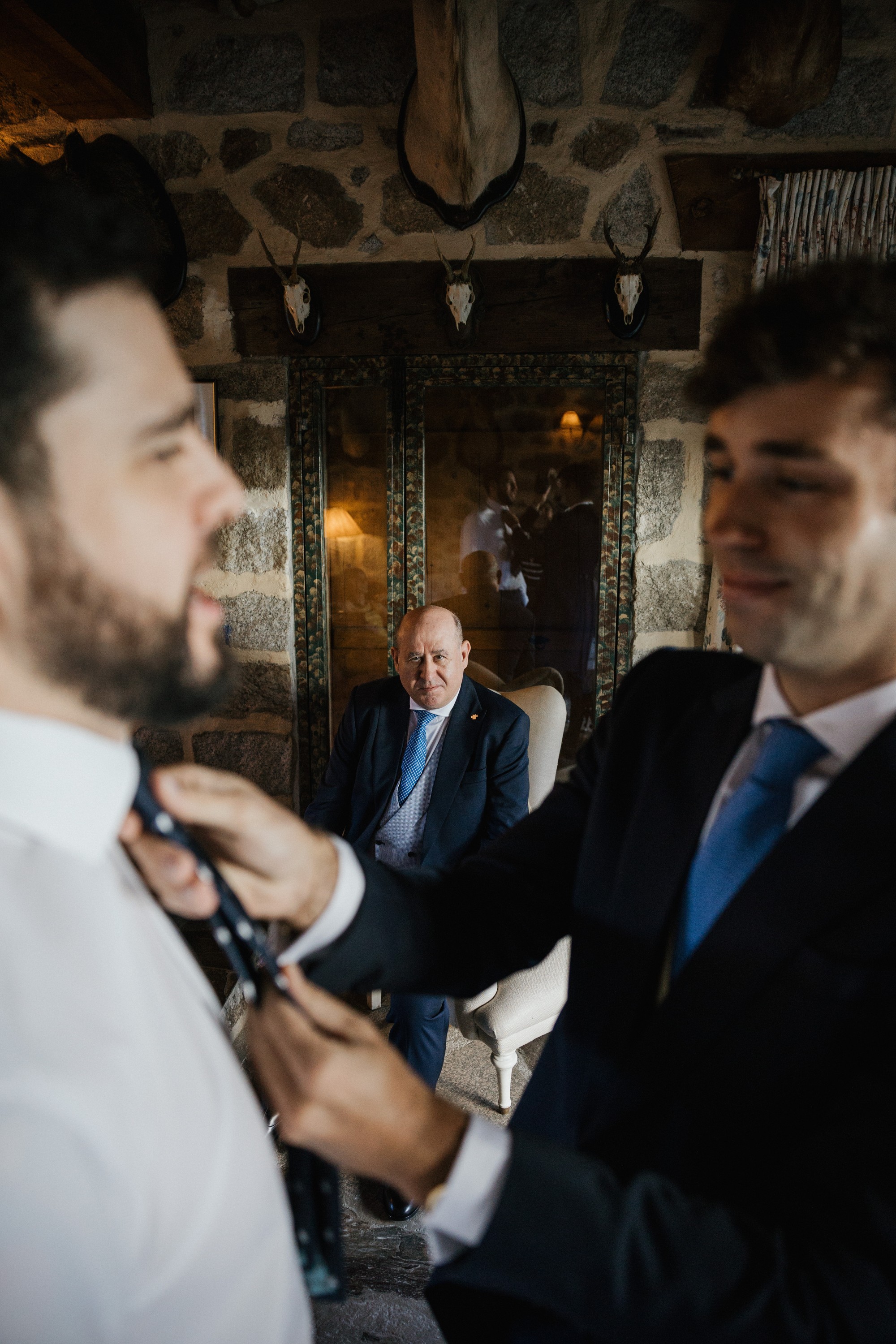 Unas manos anudan la corbata del novio mientras un invitado los observa desde el fondo