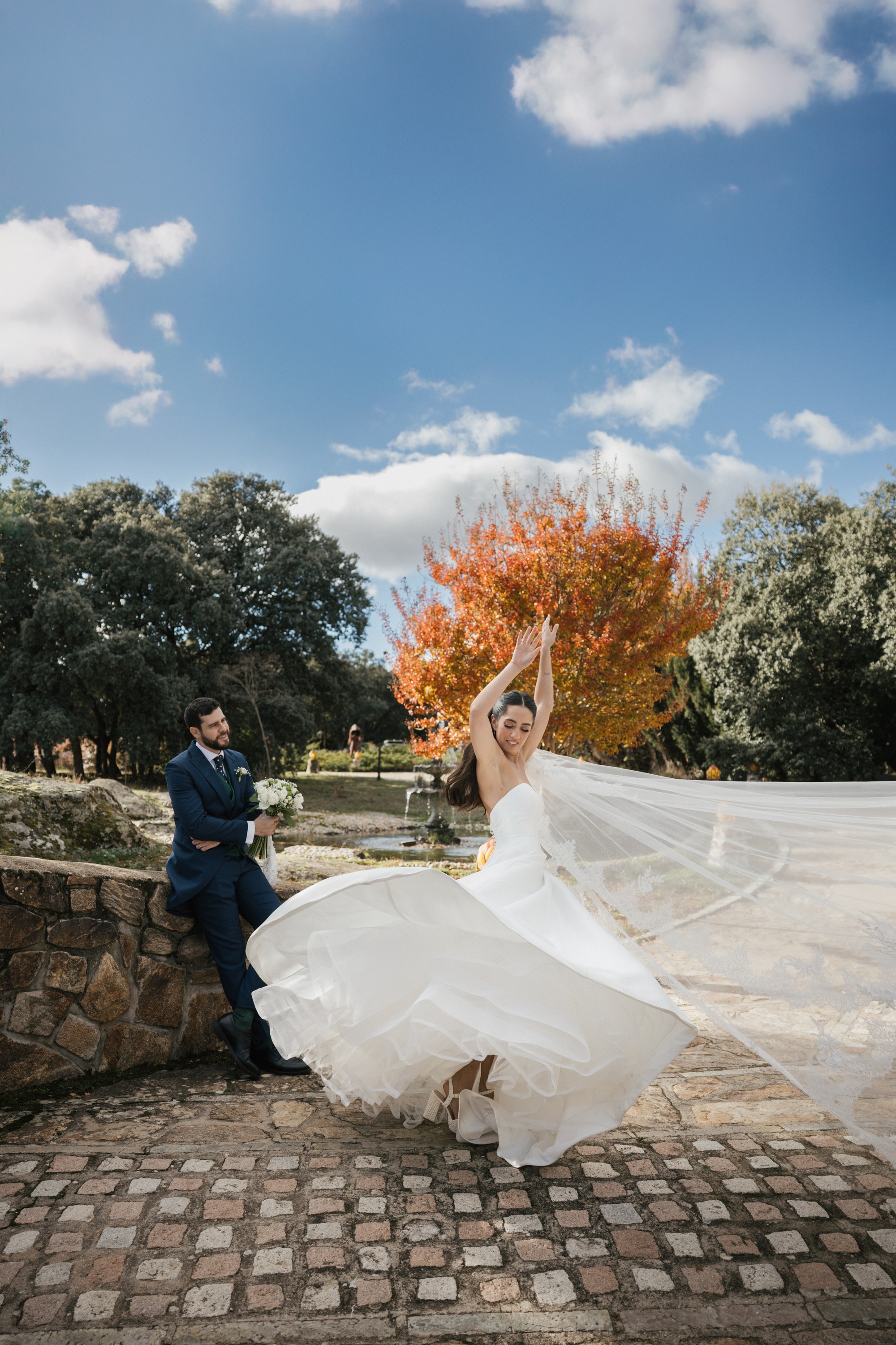 La novia gira con el velo desplegado junto a un árbol de otoño mientras el novio la observa sentado en el muro