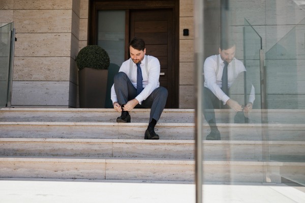 The groom ties his shoes on the front steps, mirrored in the glass panel