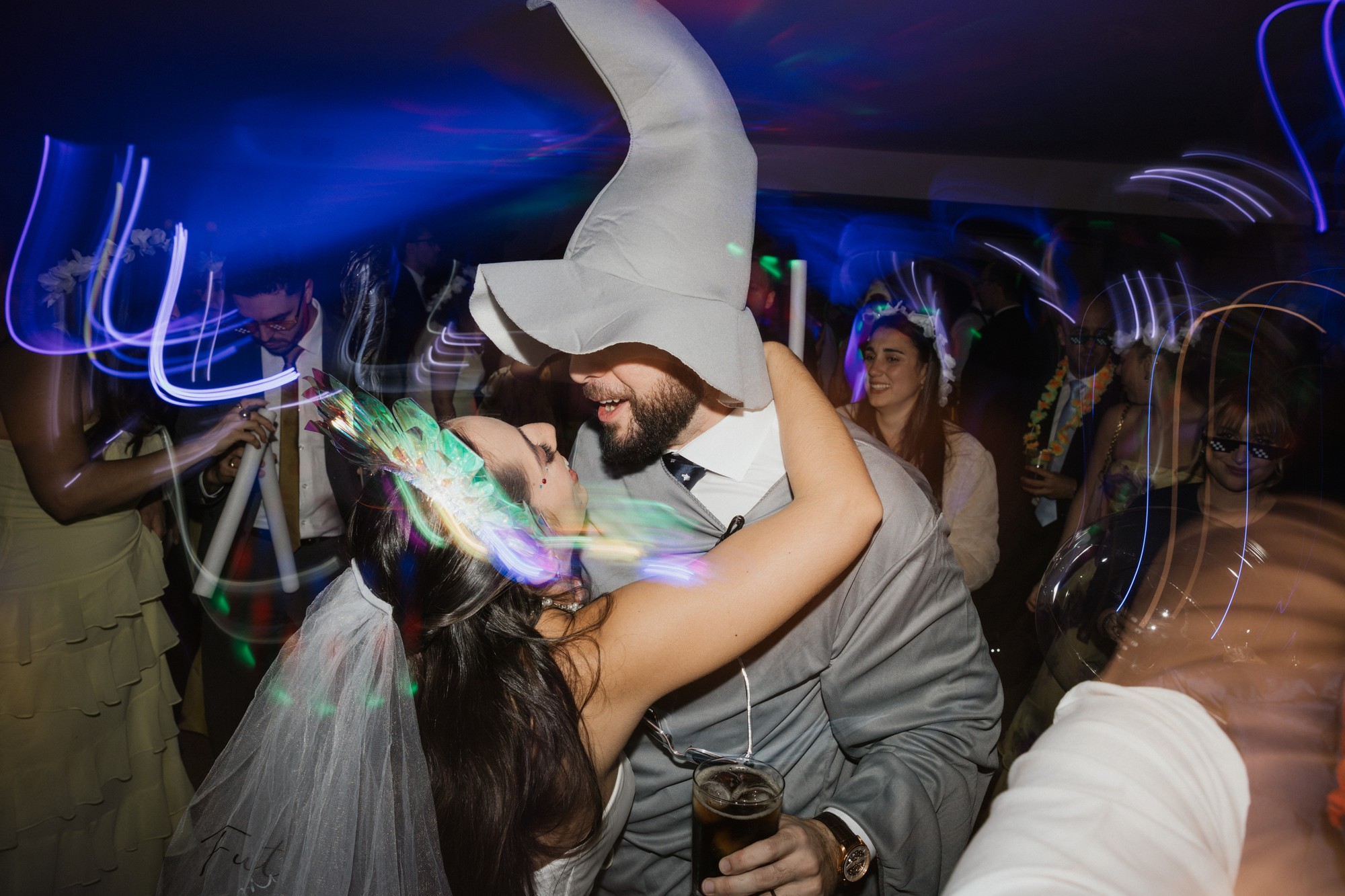 El novio baila con un sombrero de espuma mientras la novia lo abraza entre las luces de la fiesta