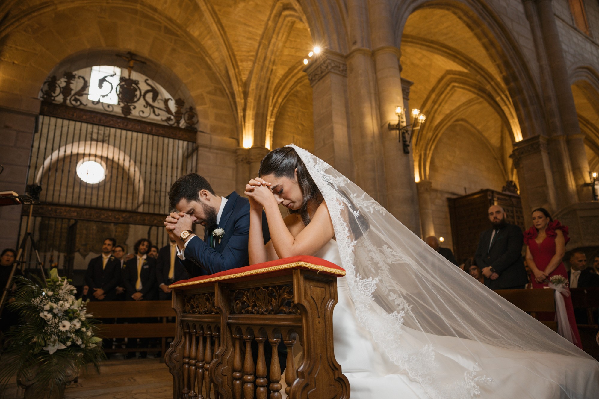 Los novios se arrodillan en el reclinatorio con las manos juntas y el velo extendido por el suelo