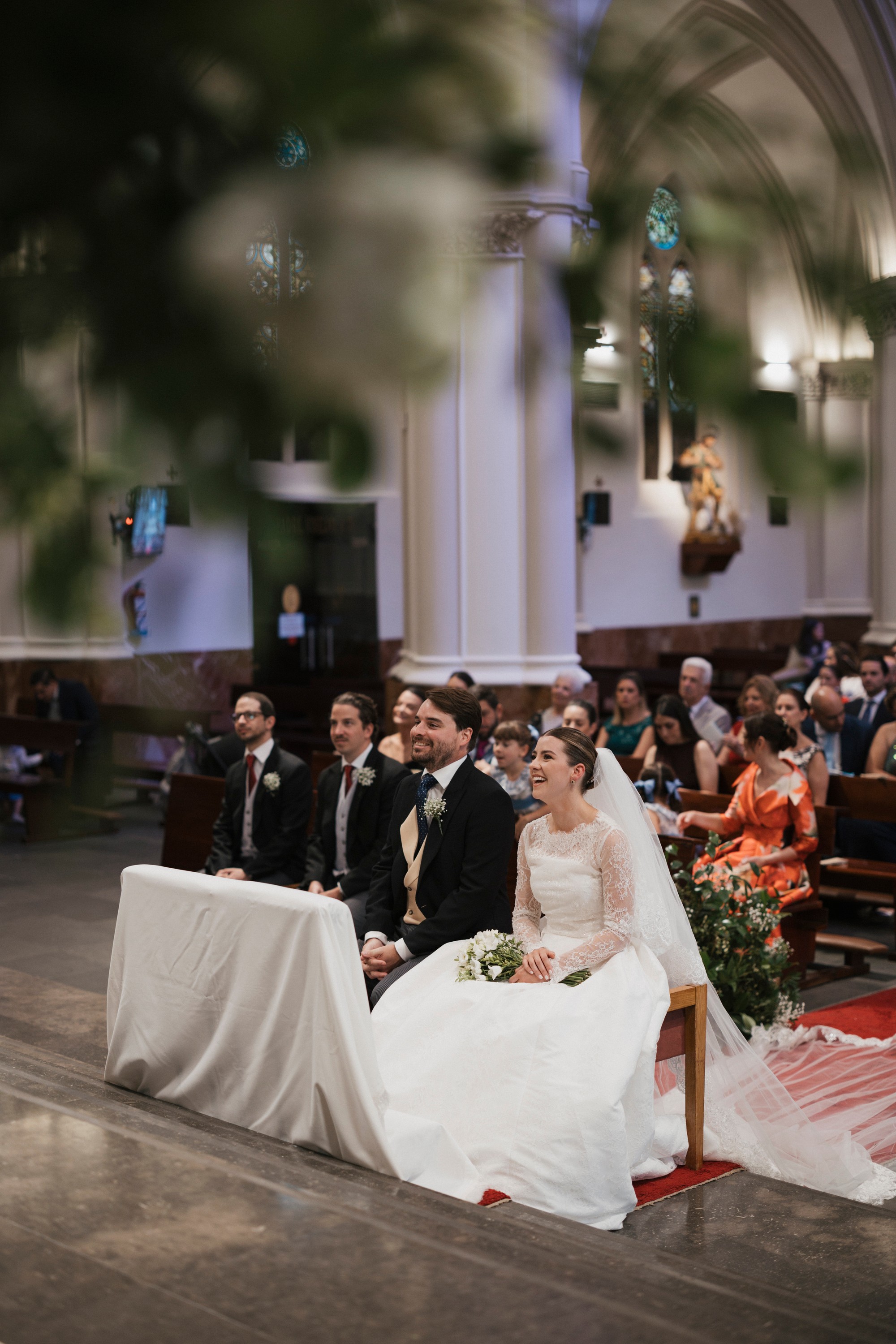 Los novios se ríen sentados durante la ceremonia, entre el verde de la decoración