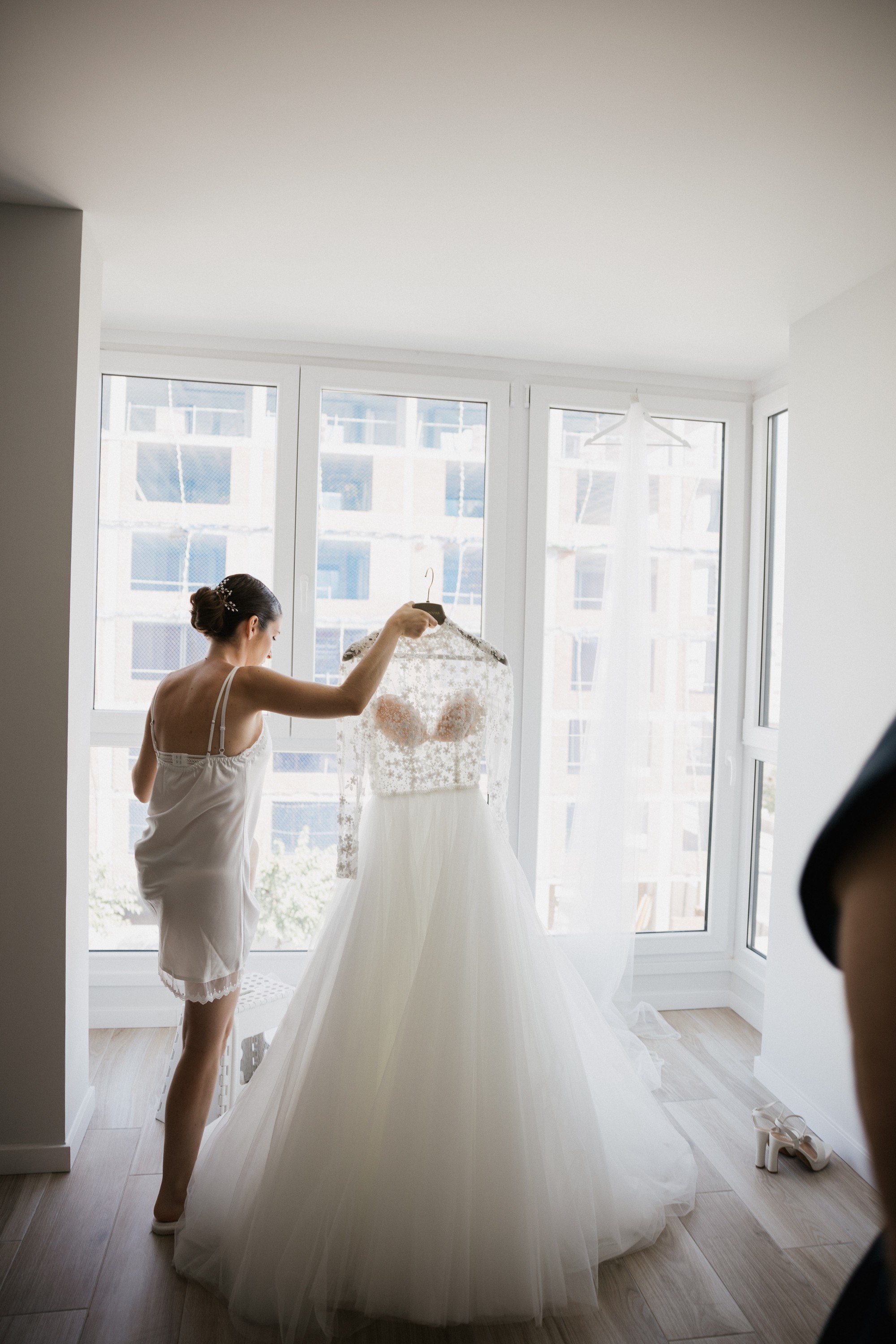 La novia sostiene en alto su vestido de encaje a contraluz de los ventanales