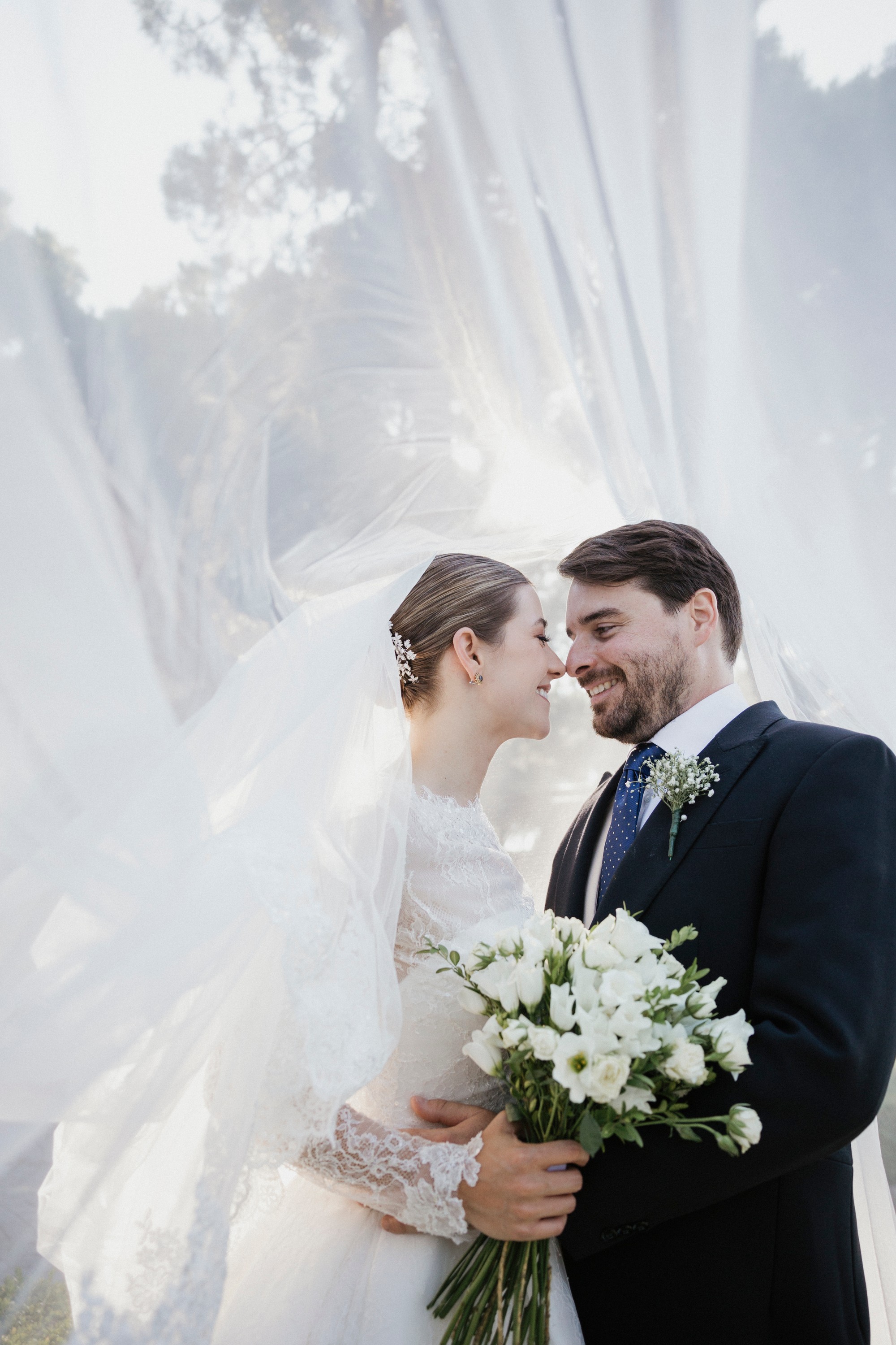 Los novios se rozan la nariz bajo el velo que vuela a su alrededor