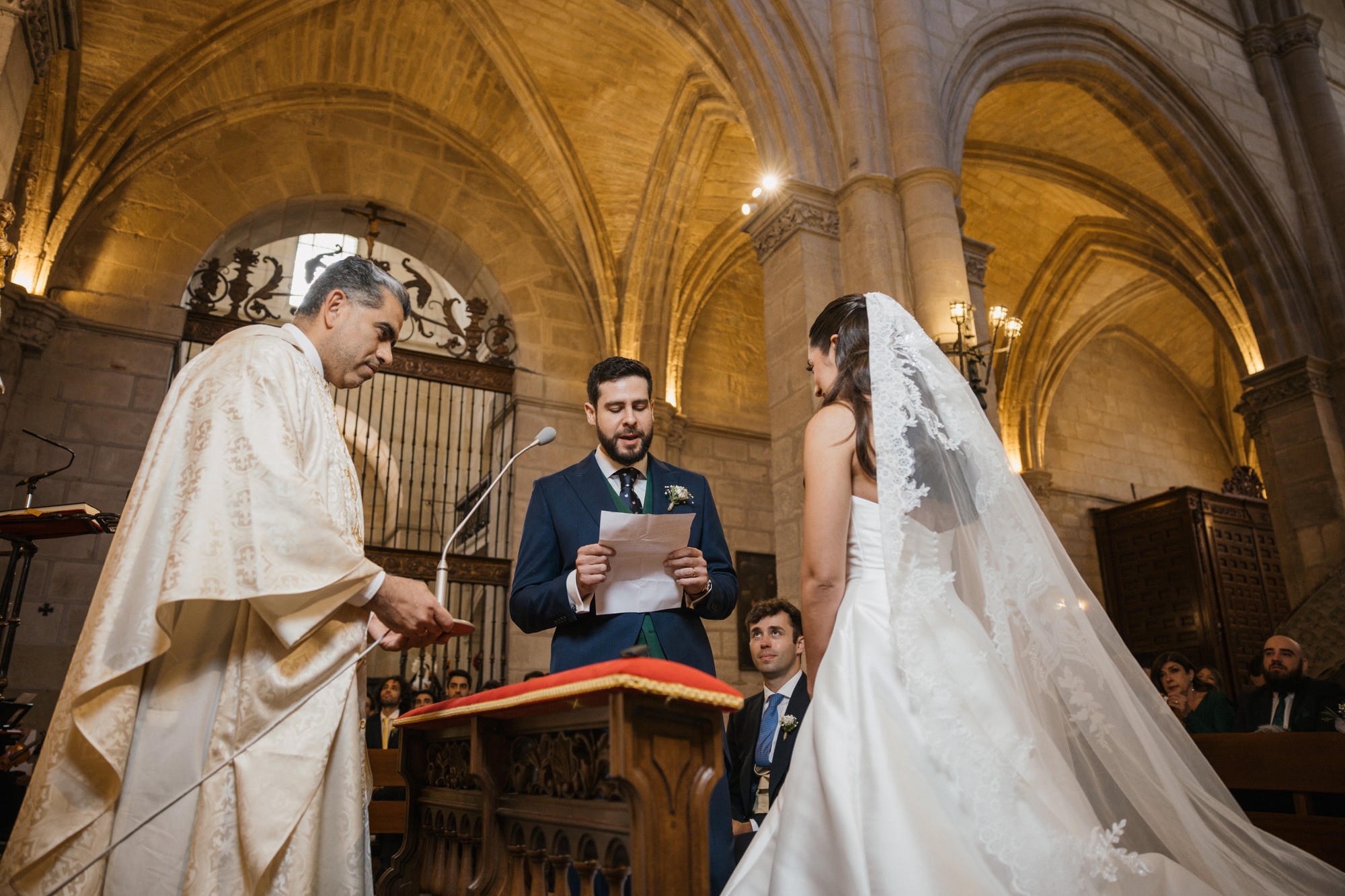 El novio lee unas palabras en el atril con la novia frente a él y el sacerdote a un lado