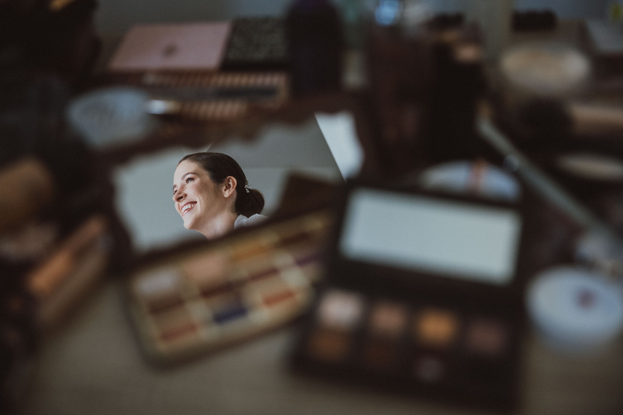 El reflejo de la novia sonriendo asoma en un espejo entre las paletas de maquillaje