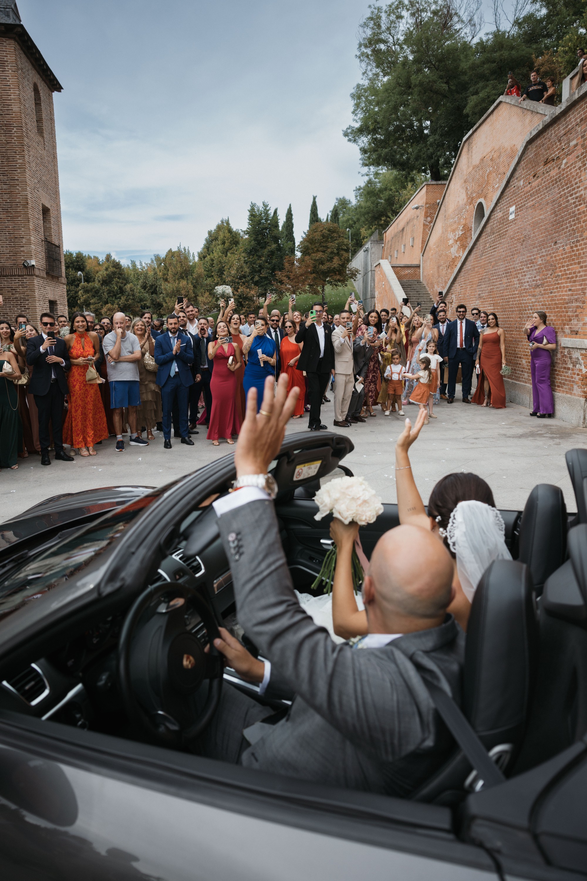 Los novios levantan los brazos desde el descapotable para despedirse de los invitados
