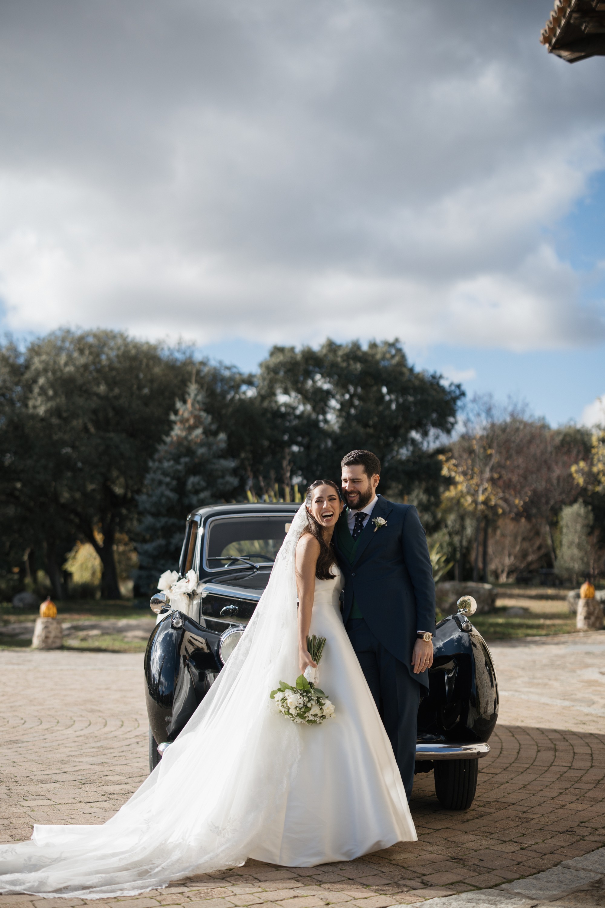 La novia ríe apoyada en el novio delante del coche clásico bajo un cielo de nubes