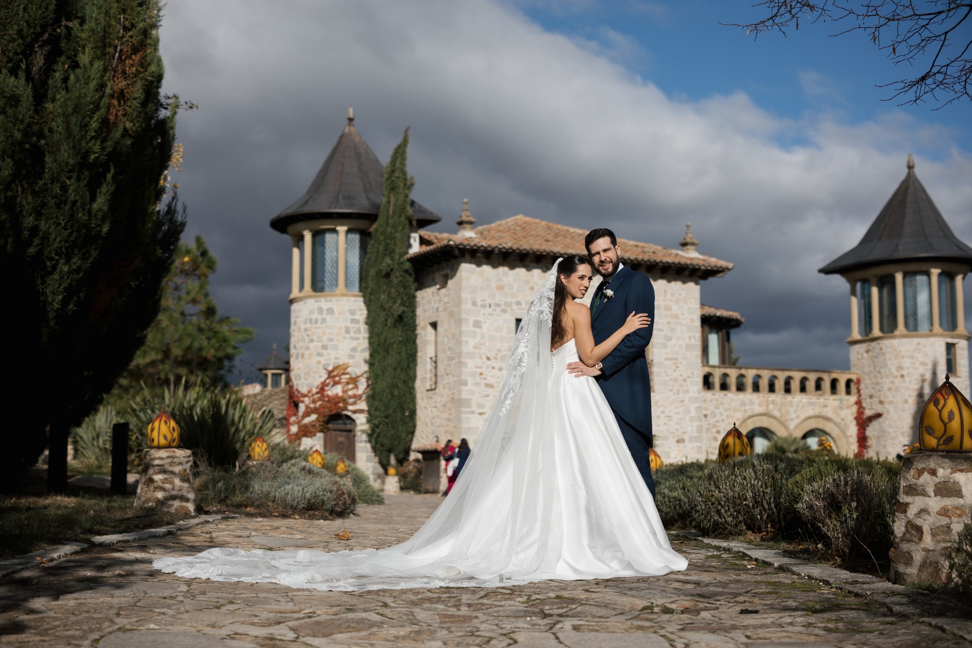 Los novios se abrazan ante la finca de piedra con torreones y un cielo de tormenta