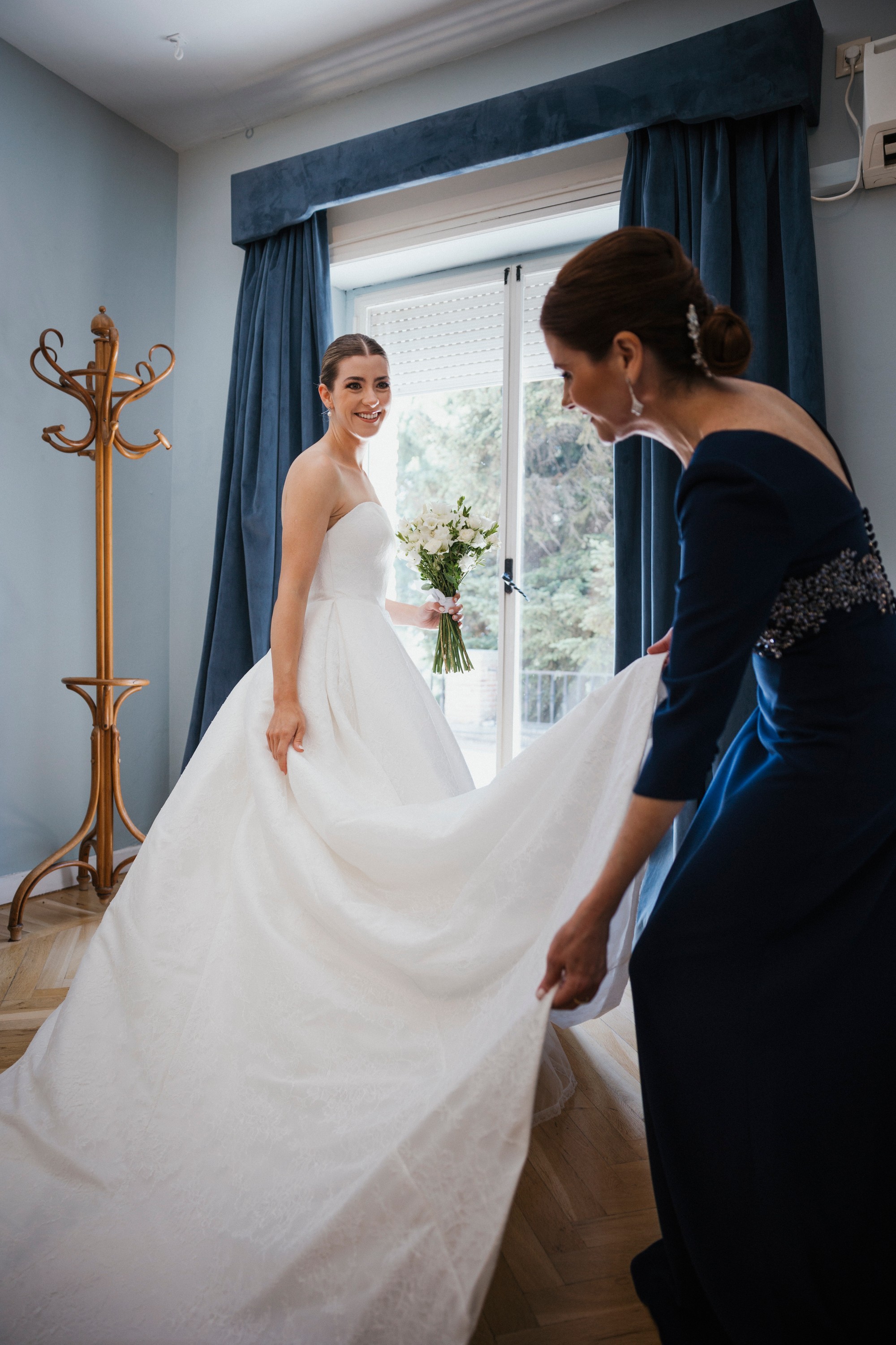 Una invitada extiende la cola del vestido mientras la novia sonríe junto a la ventana