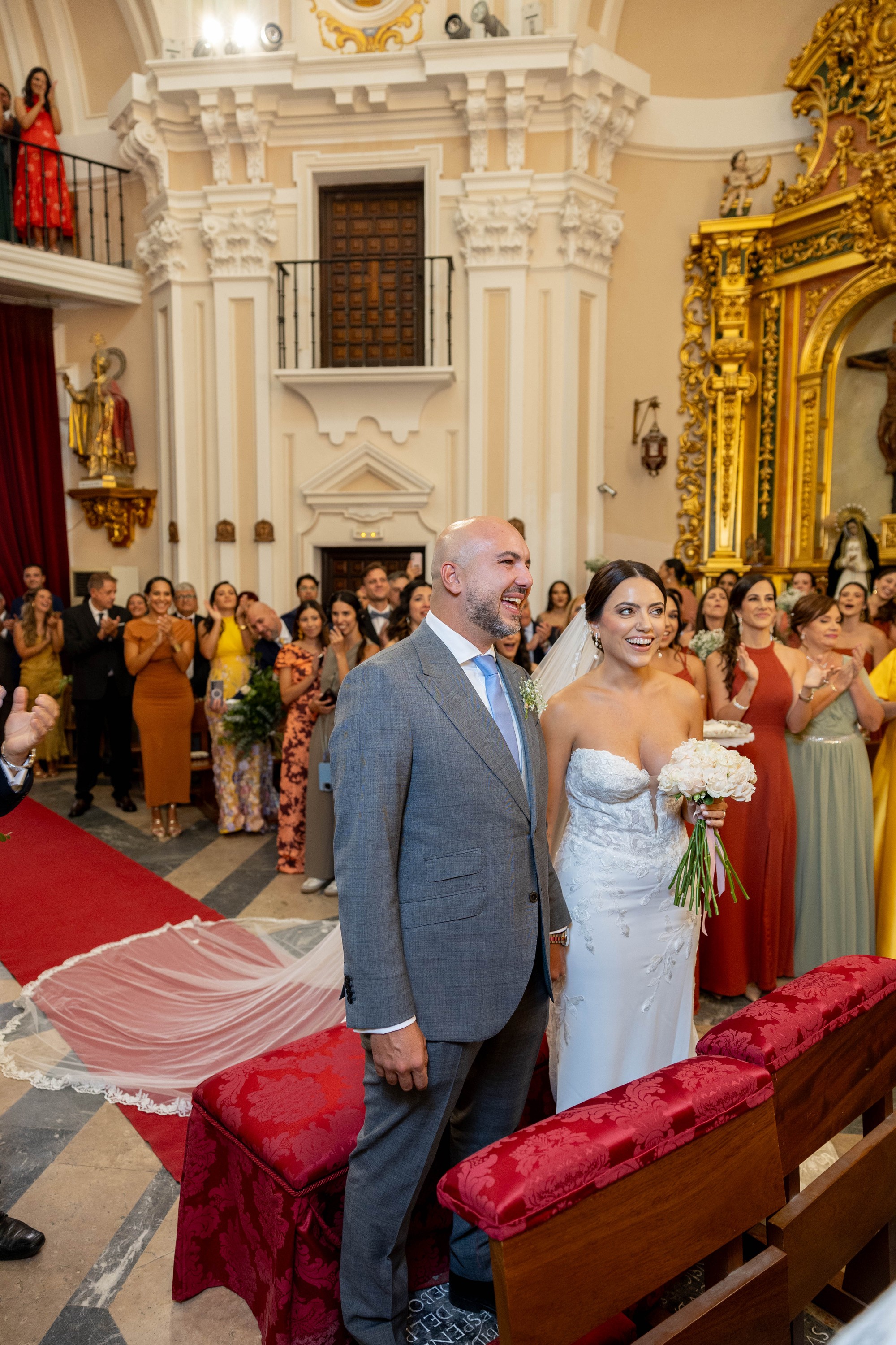Los novios sonríen de pie ante el altar con el velo desplegado sobre la alfombra roja