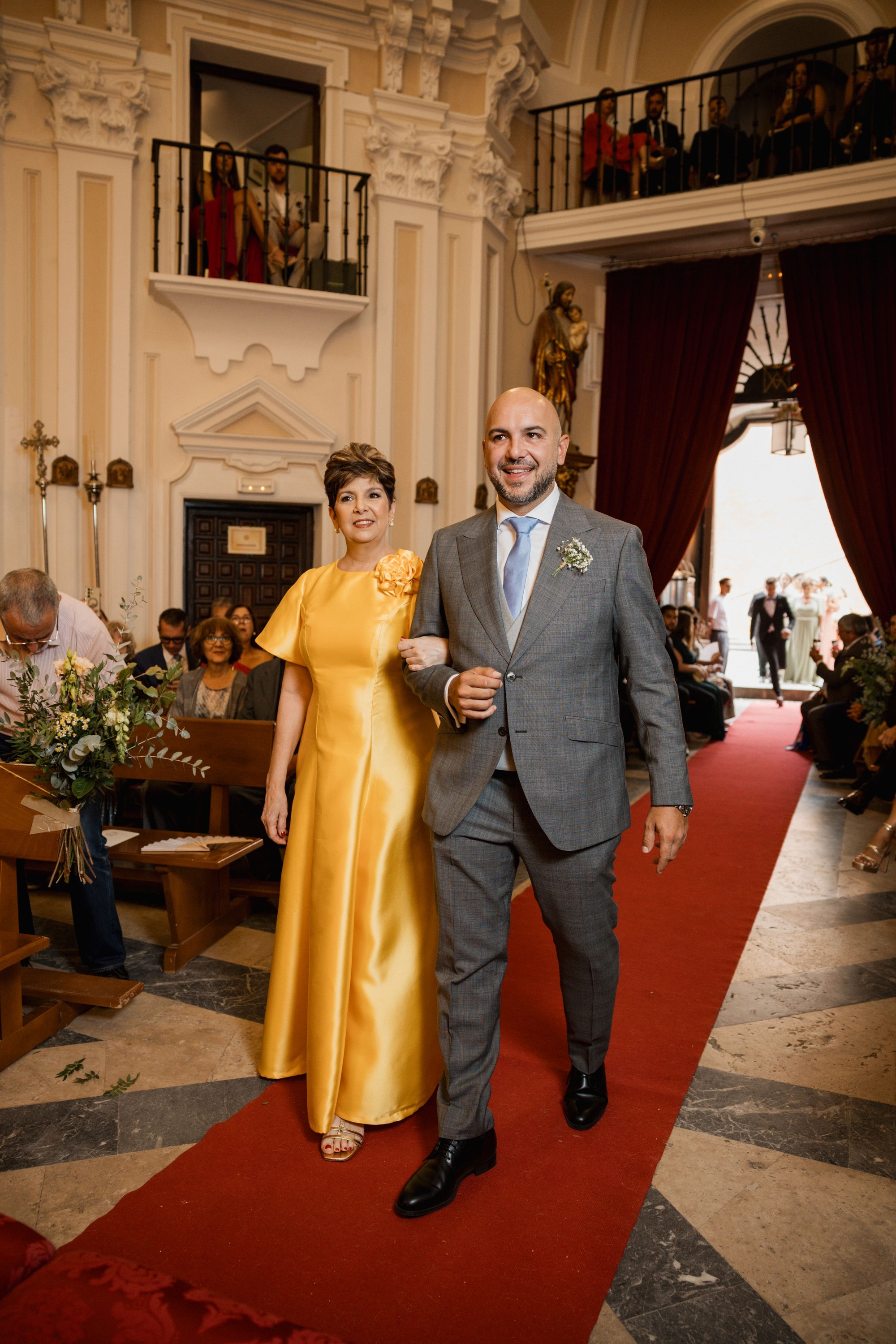 El novio recorre el pasillo de la iglesia del brazo de una invitada de vestido amarillo