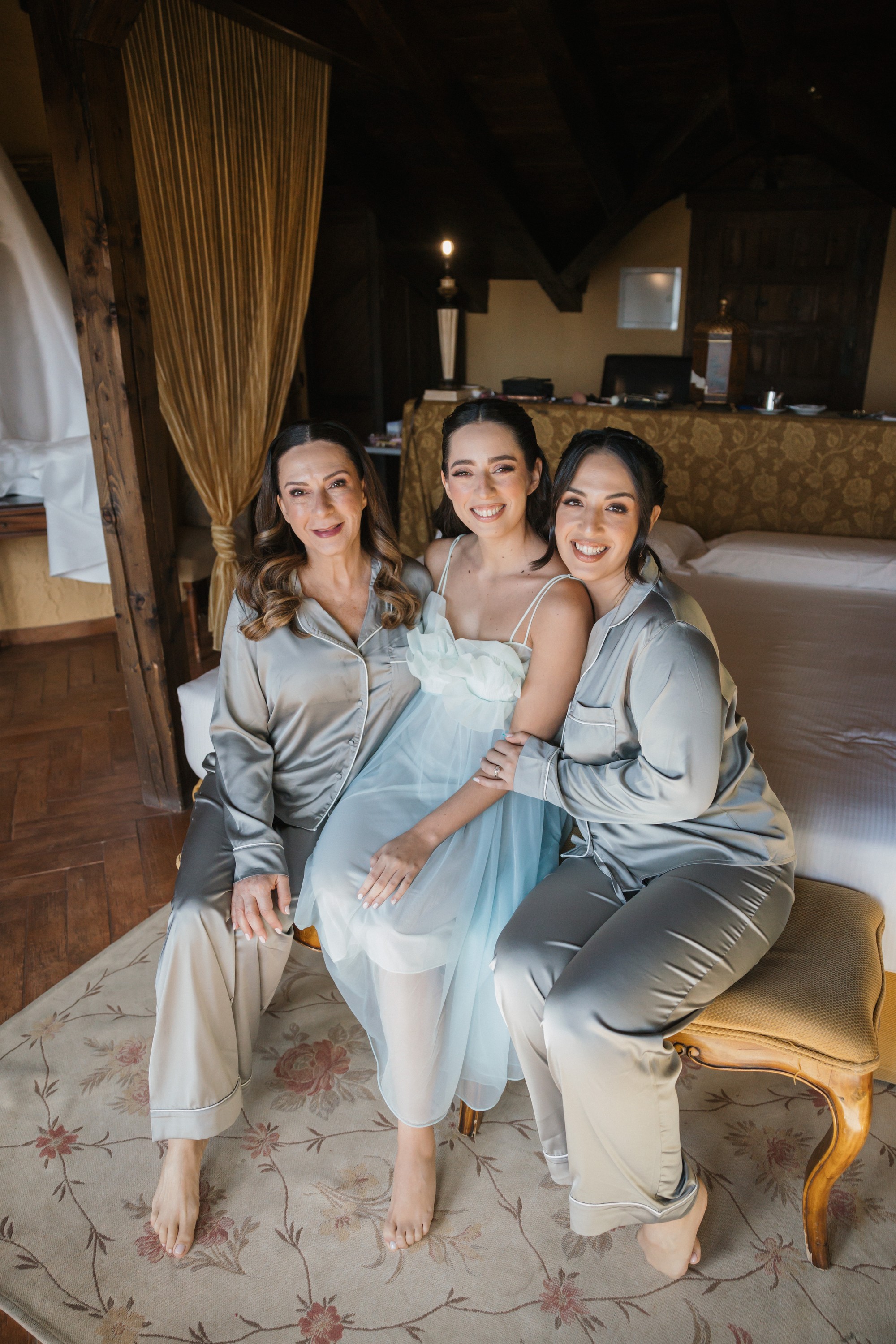 La novia posa sentada en la cama con dos invitadas, las tres en pijama de seda