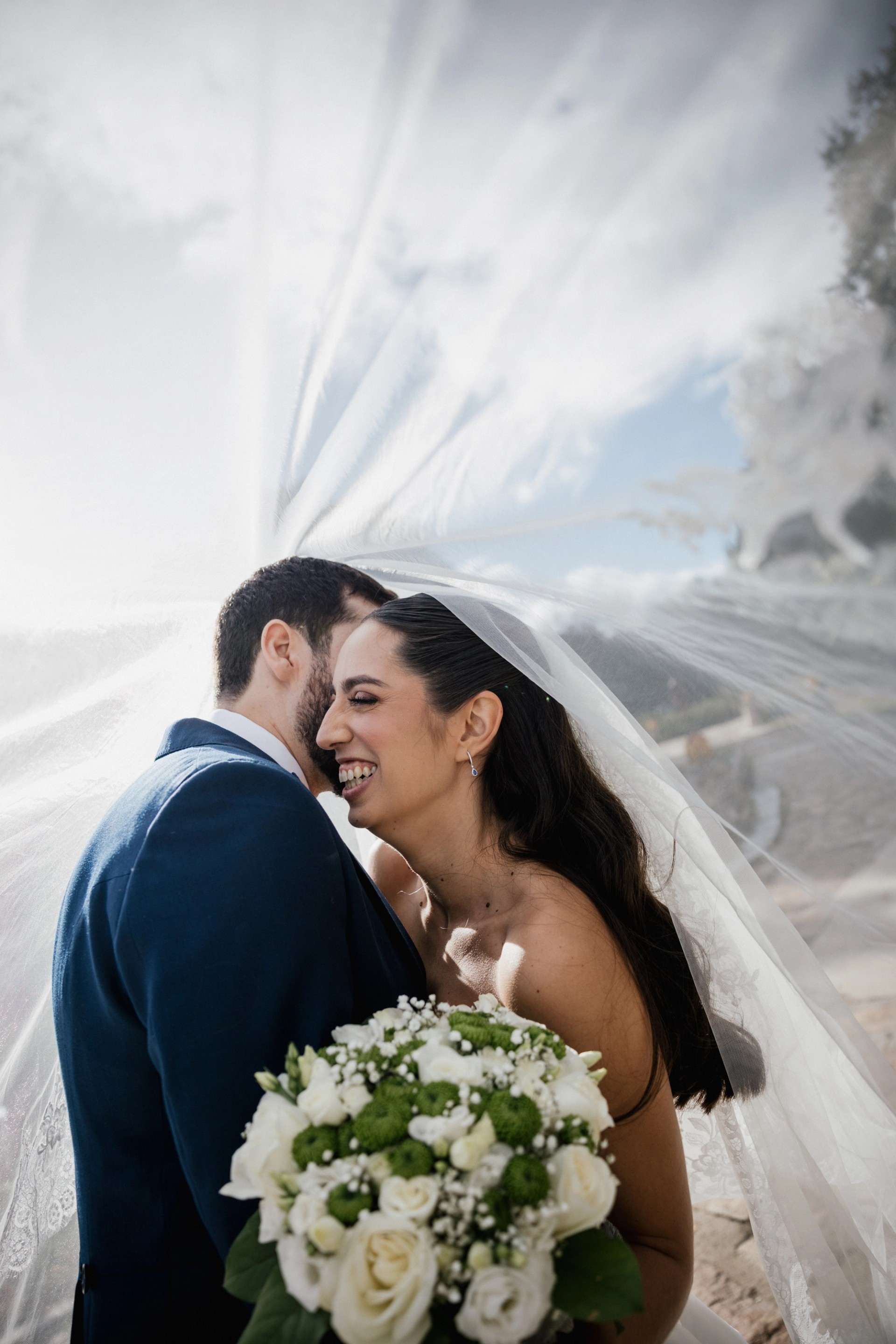 Los novios se refugian bajo el velo y la novia ríe con el ramo entre las manos