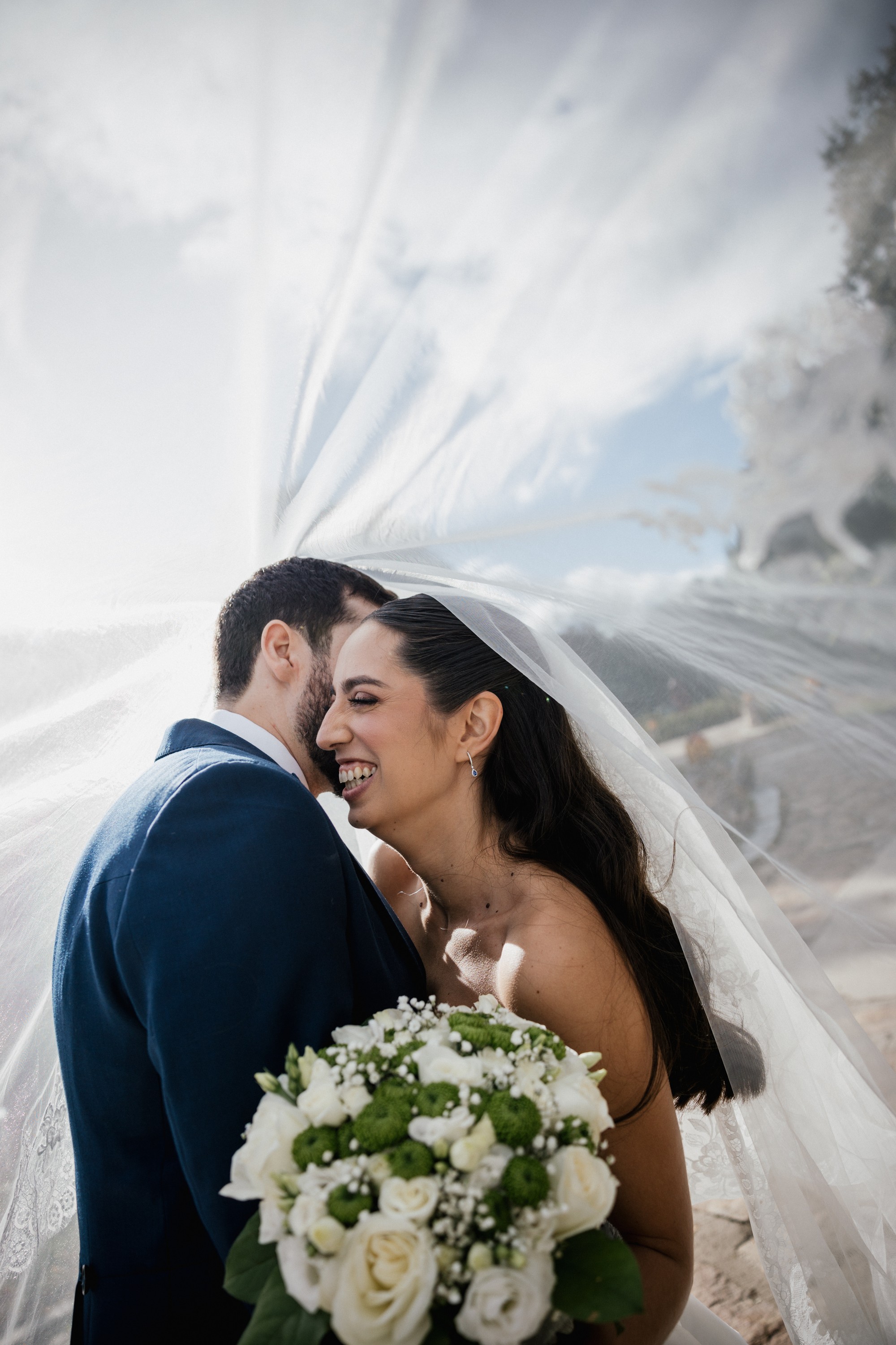 Los novios se refugian bajo el velo y la novia ríe con el ramo entre las manos