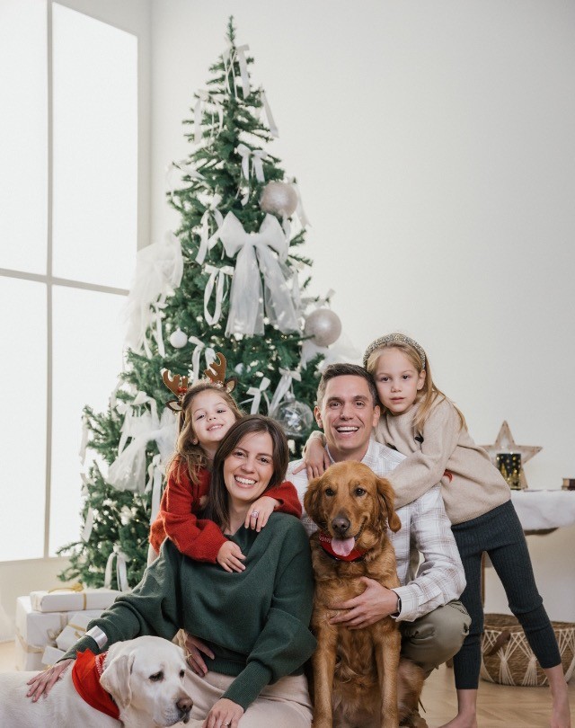 Familia con dos niñas y dos perros, un golden y un labrador, delante del árbol de Navidad con lazos blancos