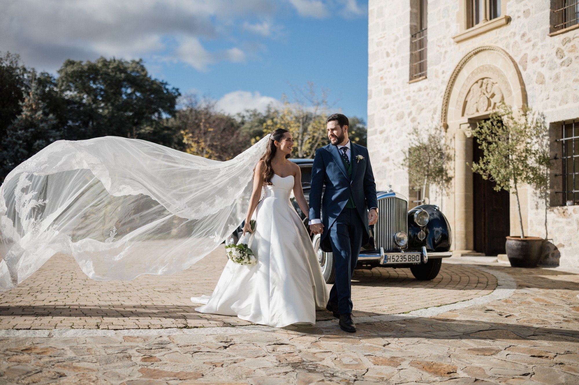 Los novios caminan de la mano junto al coche clásico con el velo volando al viento