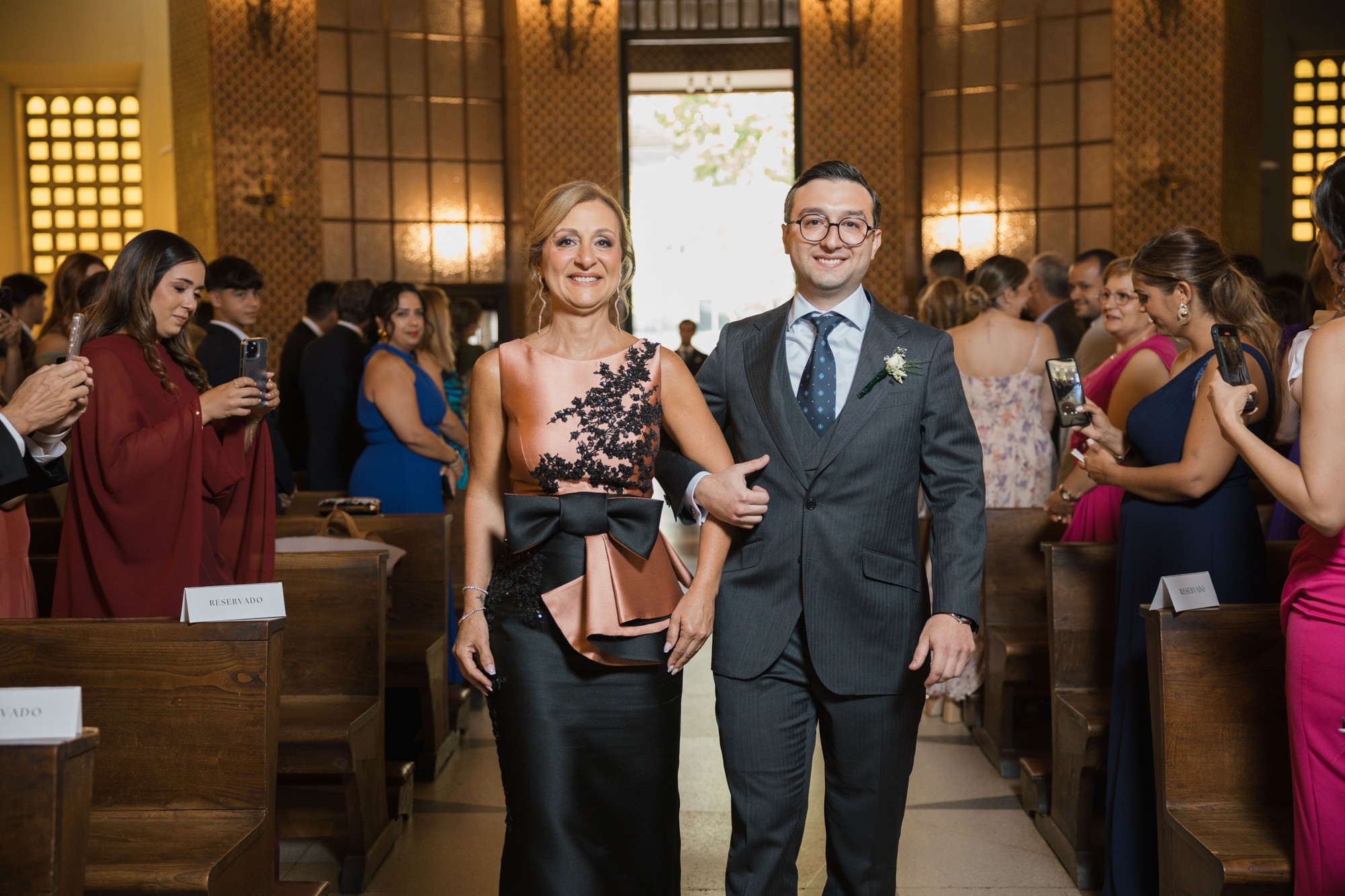 El novio recorre el pasillo de la iglesia del brazo de su acompañante mientras los invitados le graban