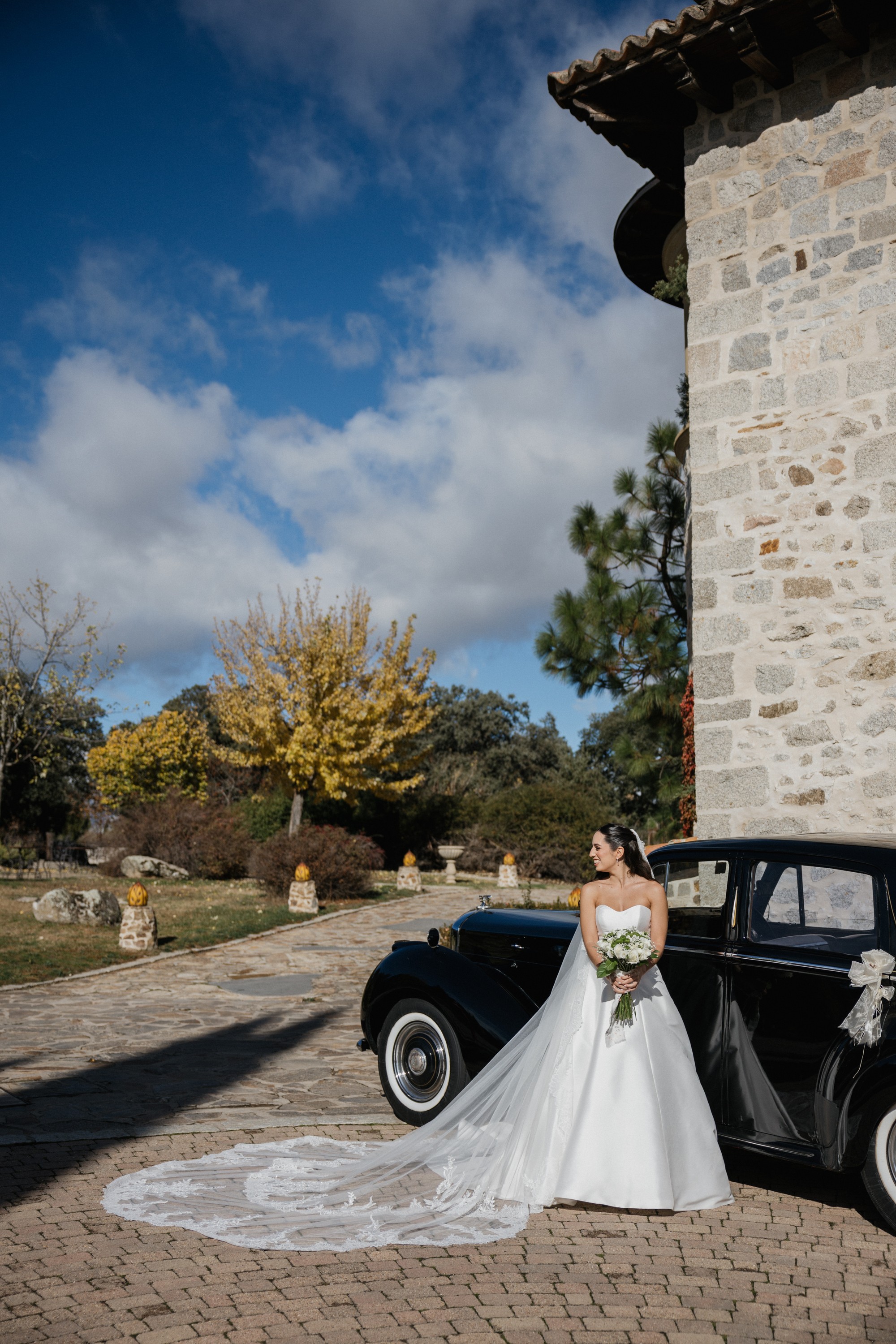 La novia espera junto al coche clásico con la cola del vestido extendida sobre el empedrado