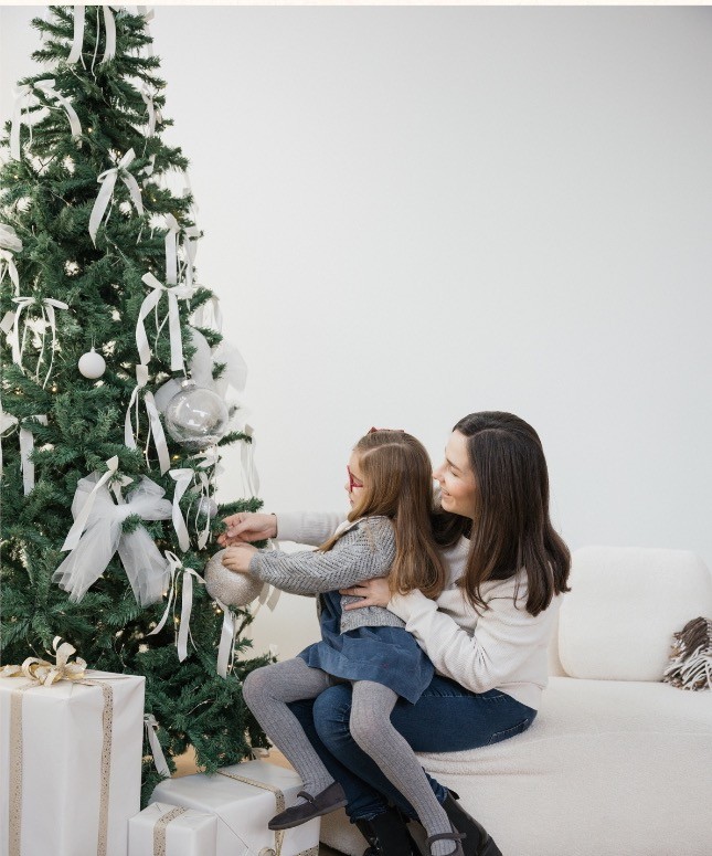 Madre e hija sentadas en un sofá blanco colocando una bola en el árbol de Navidad, con regalos envueltos en blanco y dorado