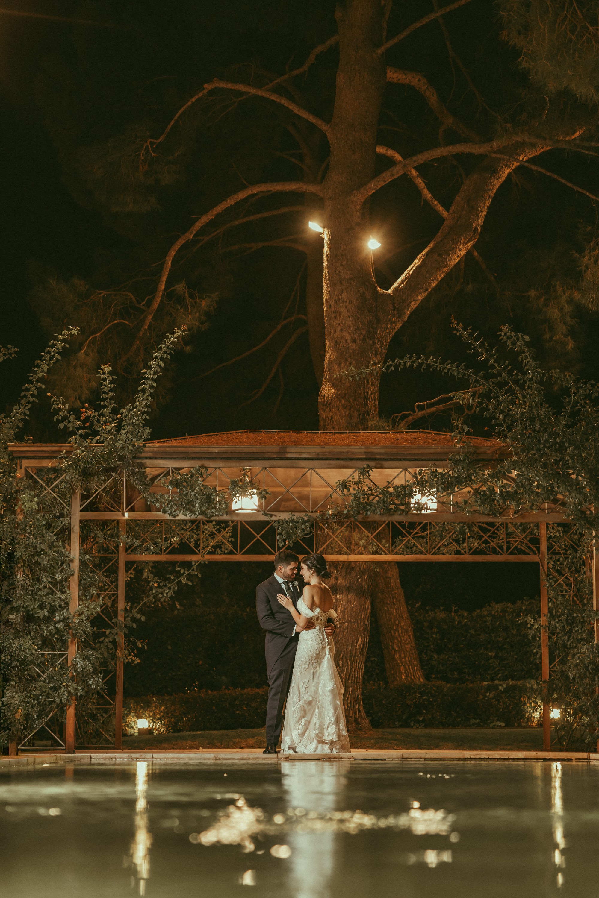 Los novios se abrazan de noche bajo la pérgola iluminada, reflejados en el agua