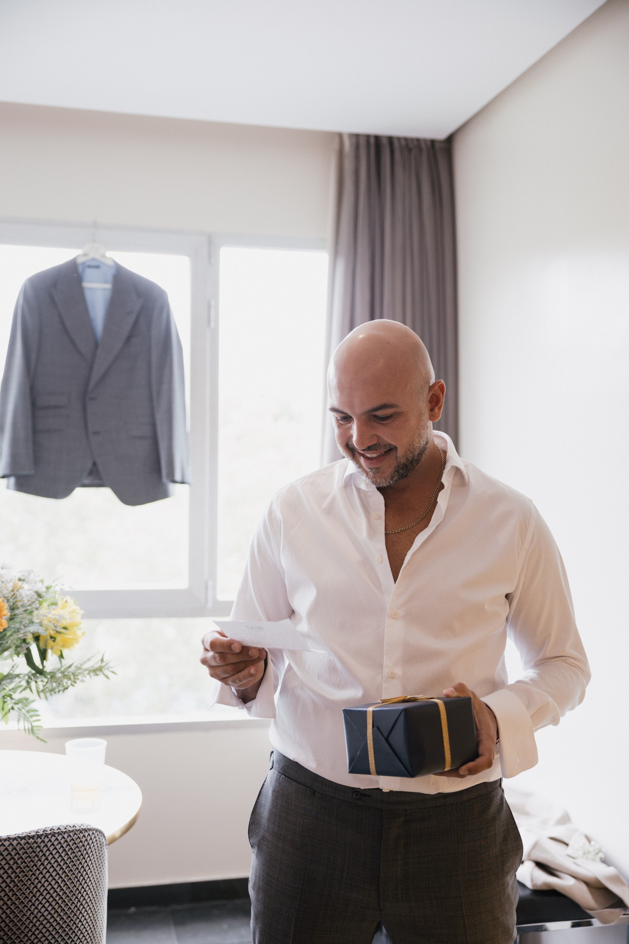 El novio lee la tarjeta que acompaña a un regalo mientras su chaqueta espera colgada en la ventana