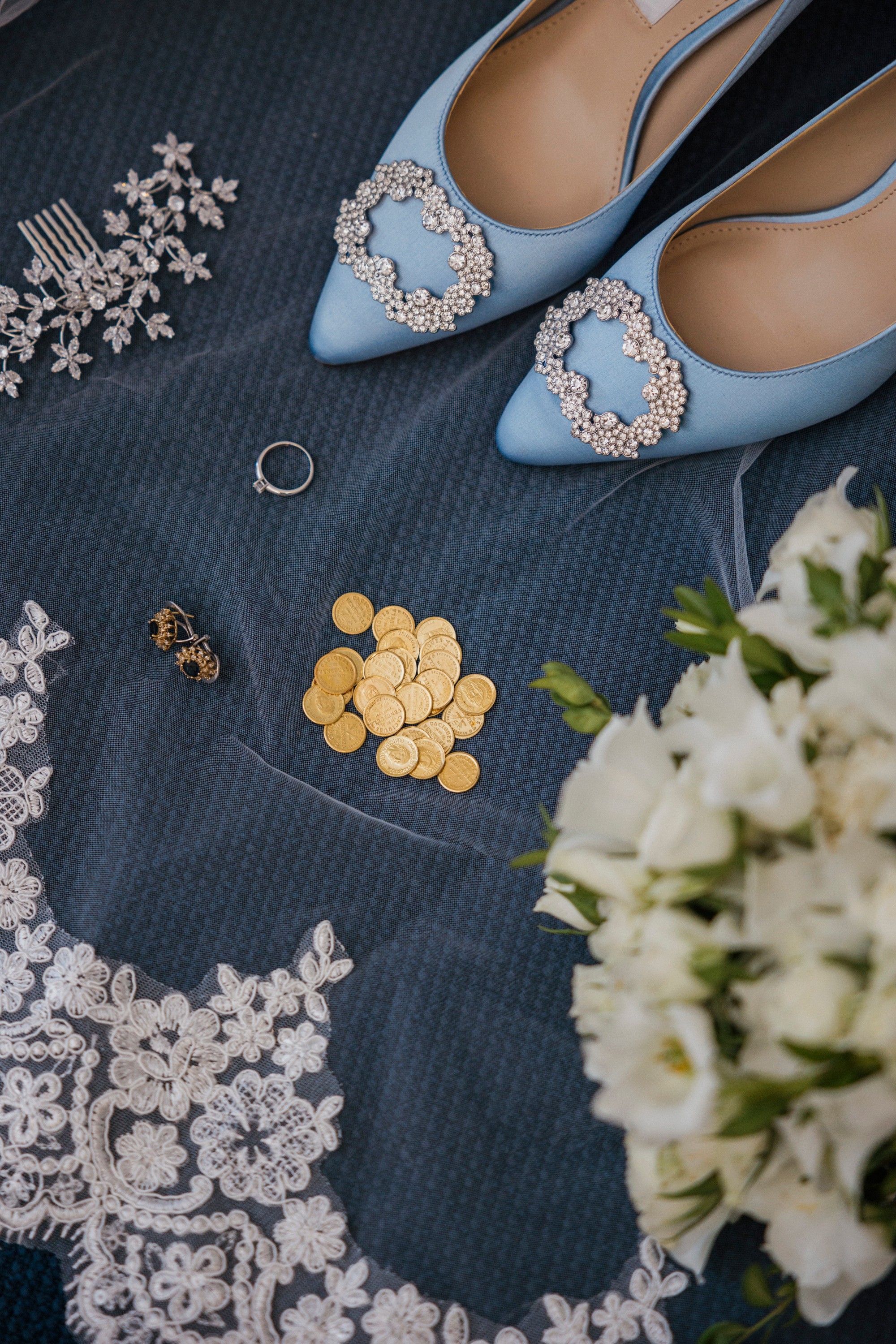 Los zapatos azules, el anillo y las arras esperan sobre el tul junto al ramo de la novia