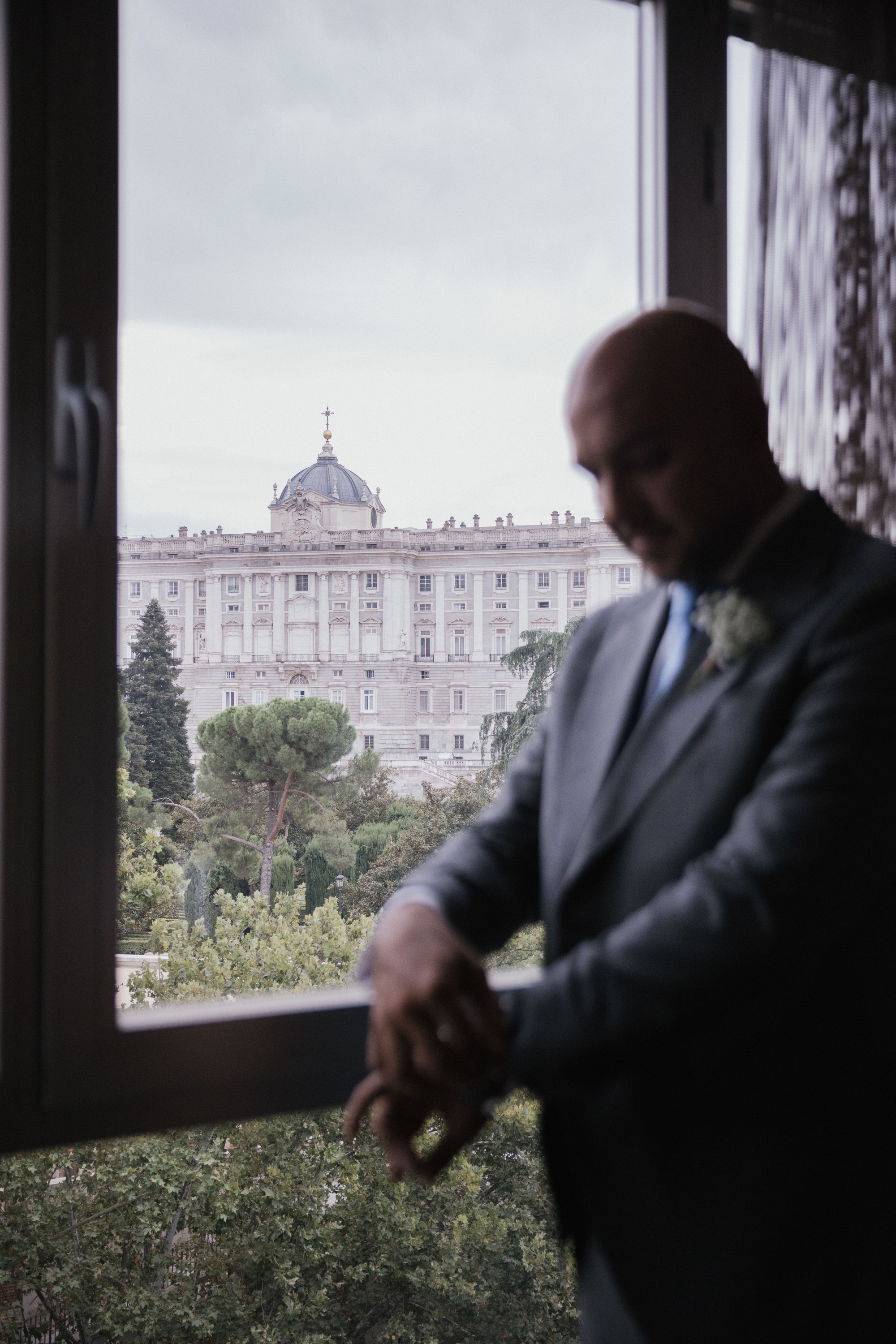 El novio se ajusta el puño de la camisa junto a la ventana, con el Palacio Real de Madrid al fondo