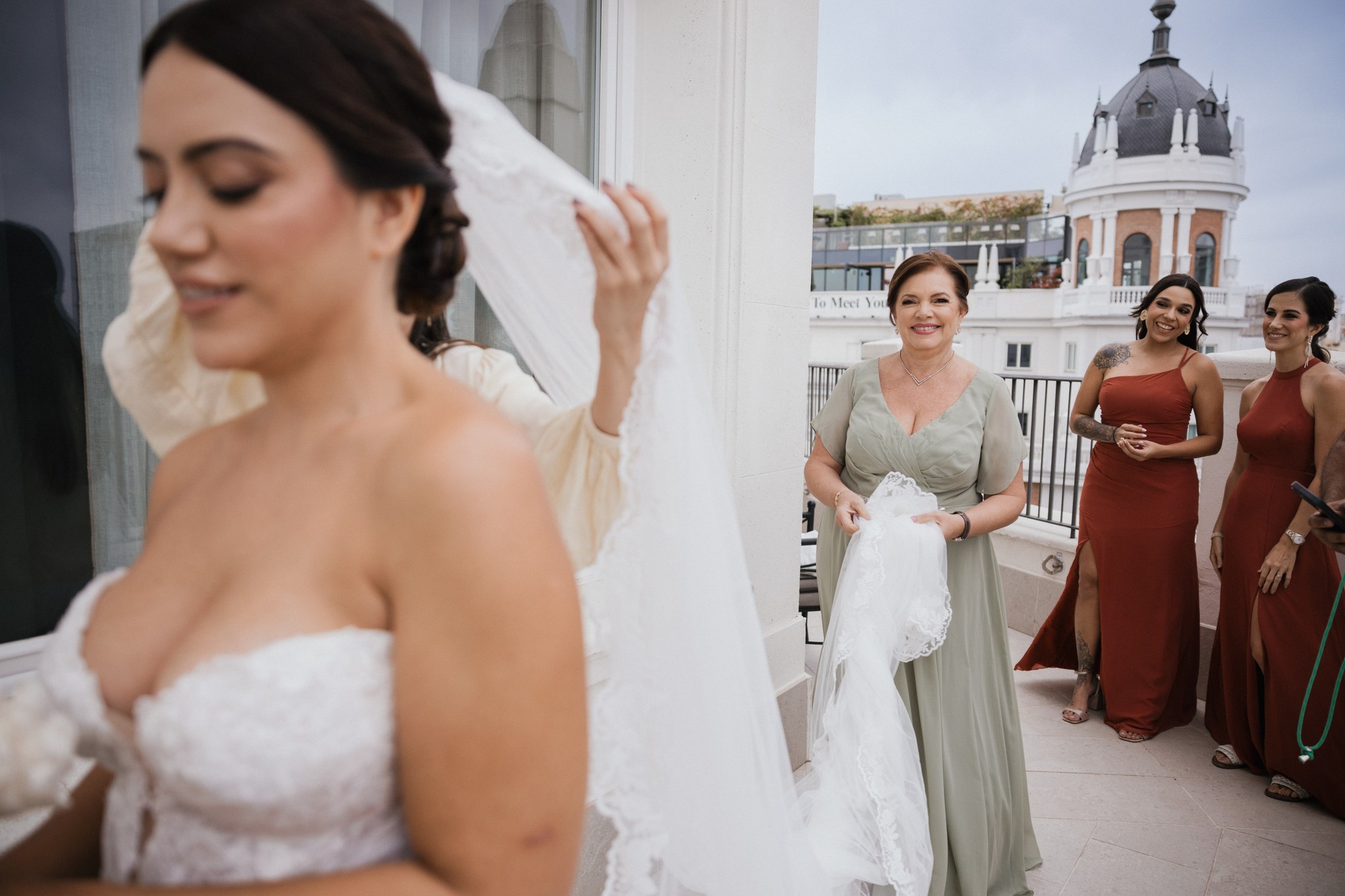 La novia se coloca el velo mientras una invitada le sostiene la cola y sus amigas esperan en la terraza