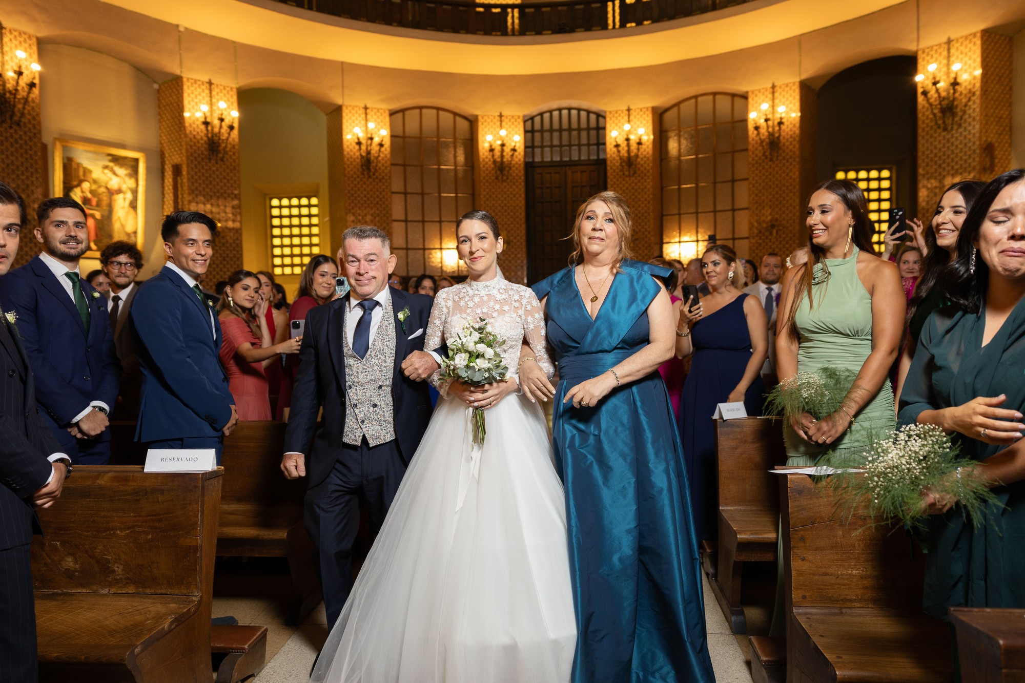 La novia avanza hacia el altar acompañada y los invitados se ponen en pie a su paso