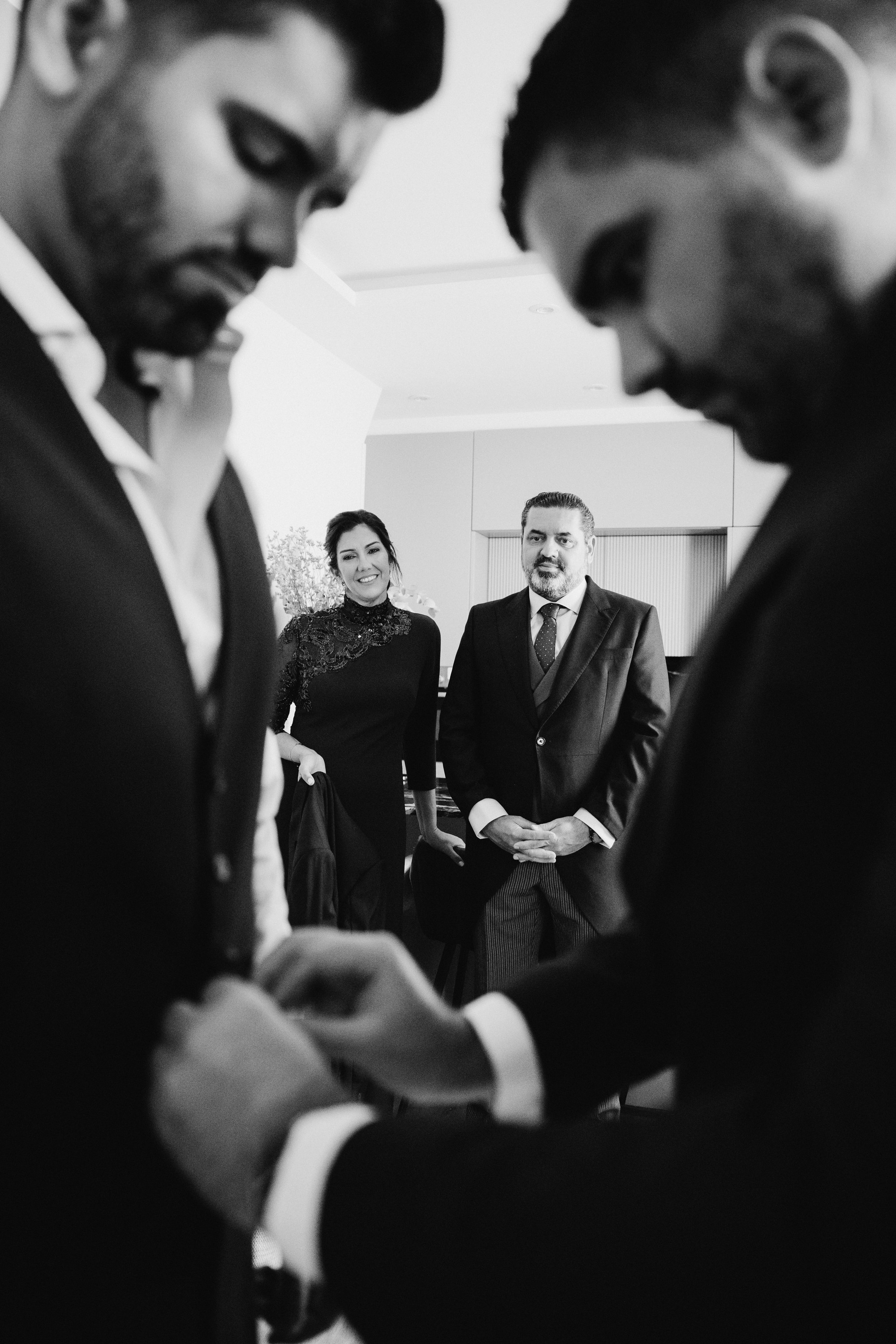 Le abrochan los gemelos al novio mientras dos invitados sonríen al fondo