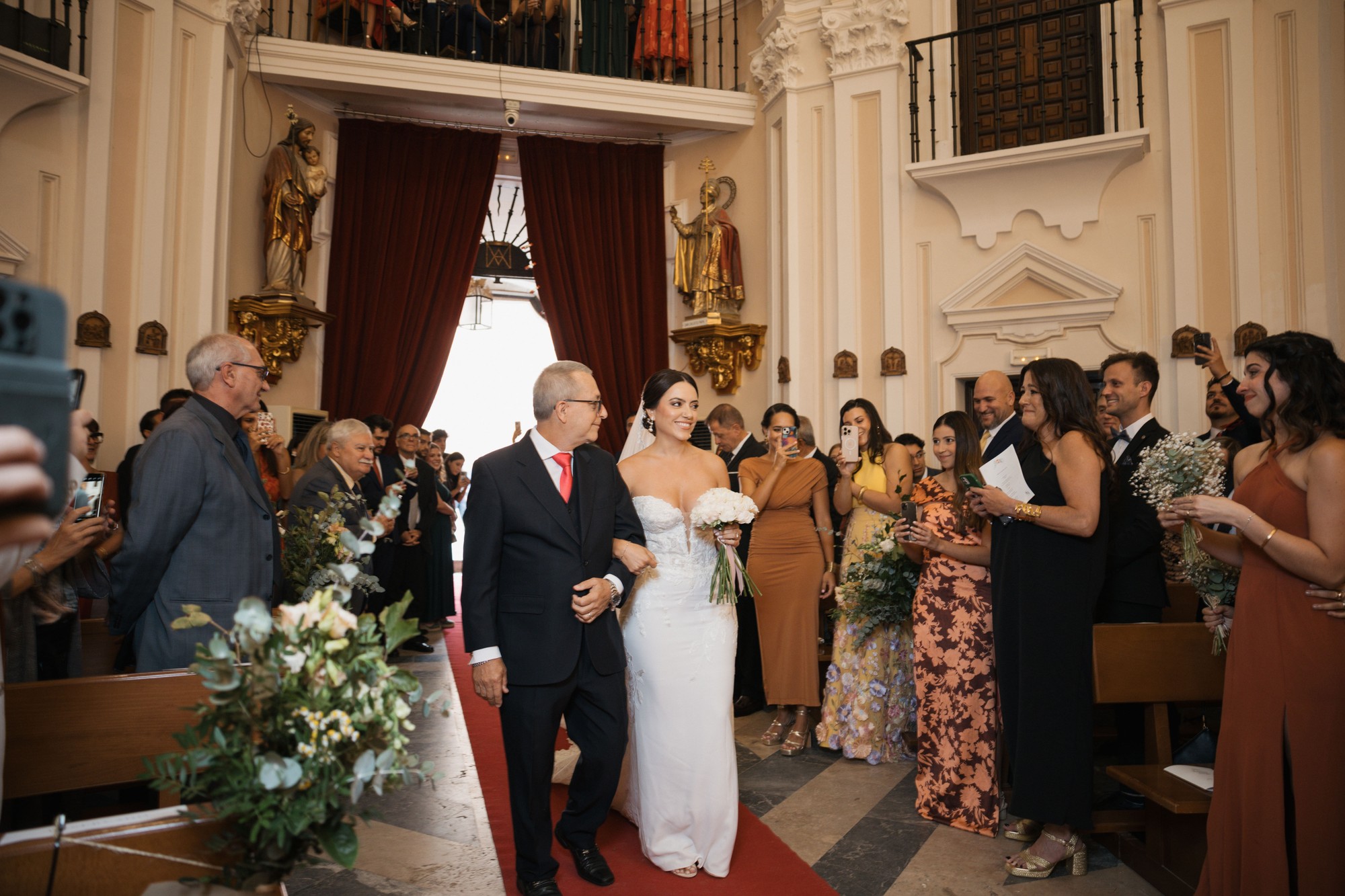 La novia entra en la iglesia del brazo de un invitado mientras los asistentes la graban con el móvil