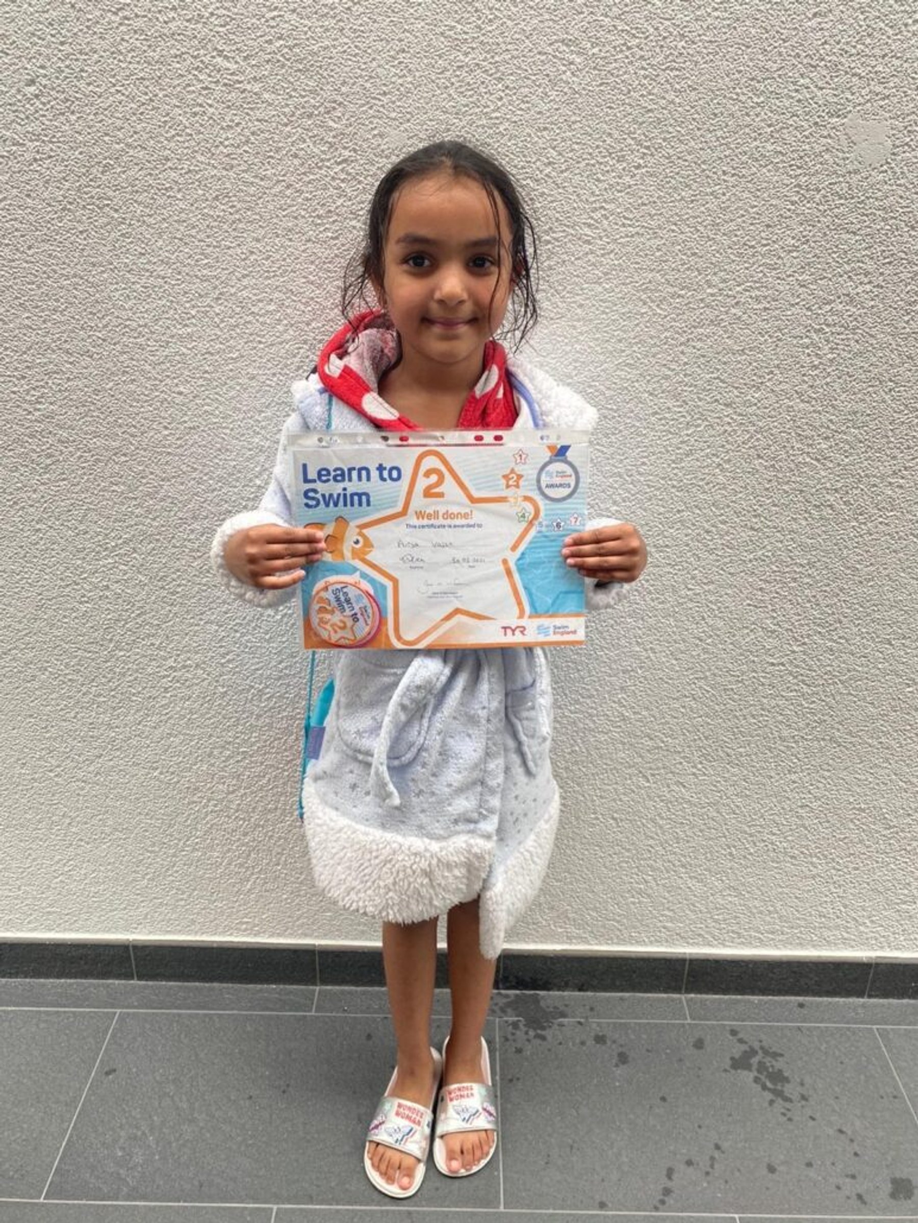 A young swimmer in a towel robe holding a Learn to Swim certificate