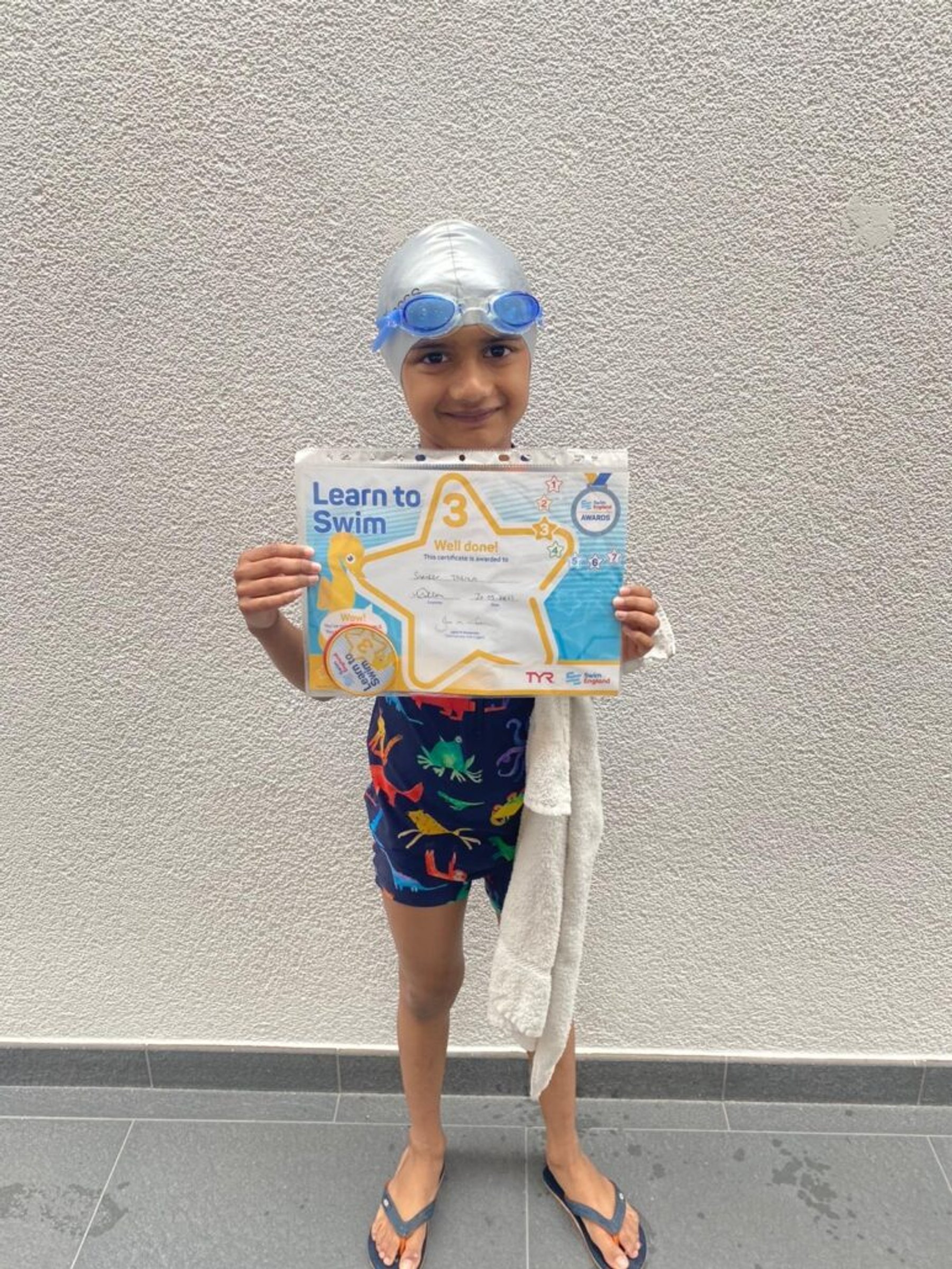A young swimmer with goggles on, holding a Learn to Swim certificate