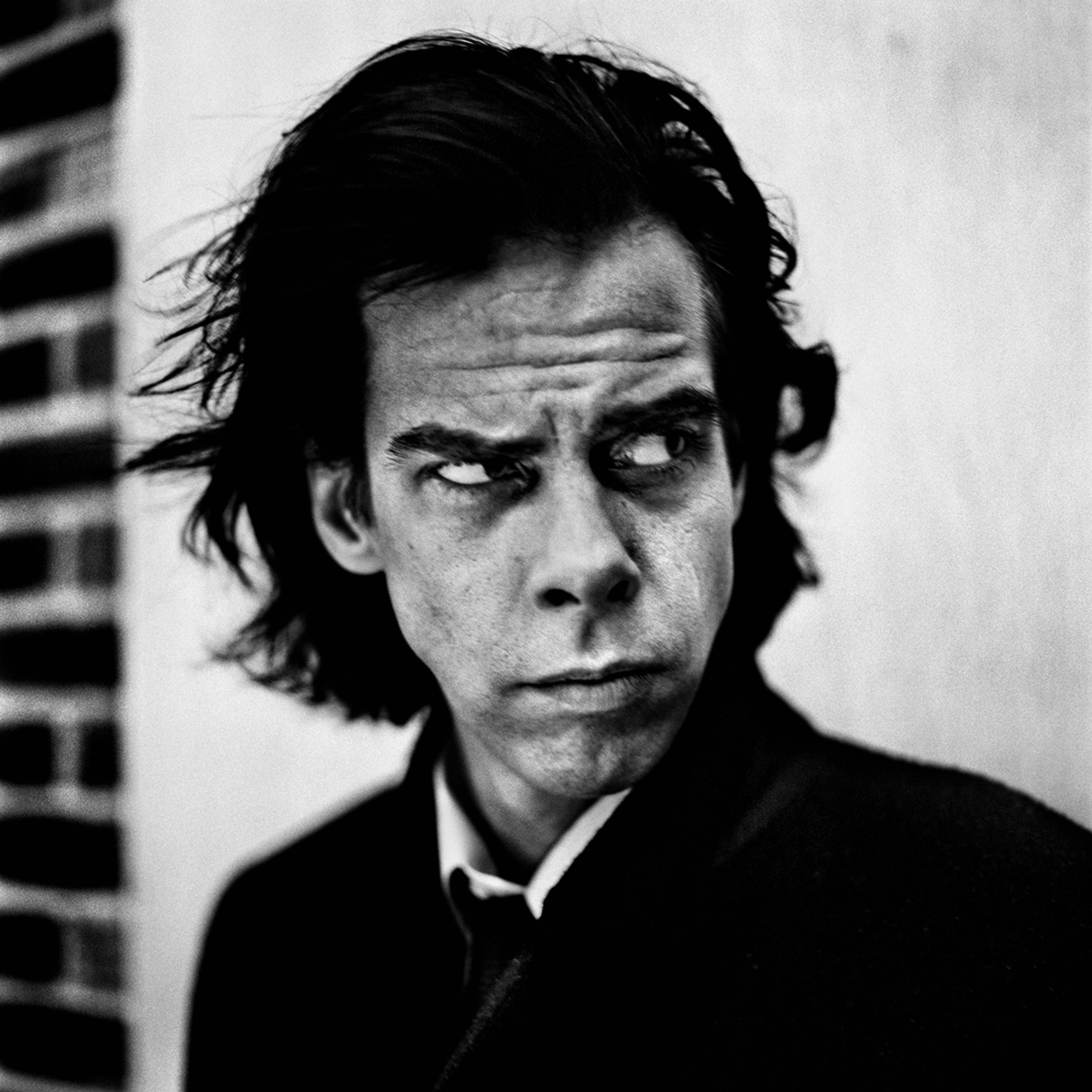 Portrait of Nick Cave, photographed in black and white by Anton Corbijn in London, 1996. Nick Cave is seen with dark, disheveled hair and a serious, contemplative look directed to the side. The background is blurred with a brick wall on the left.