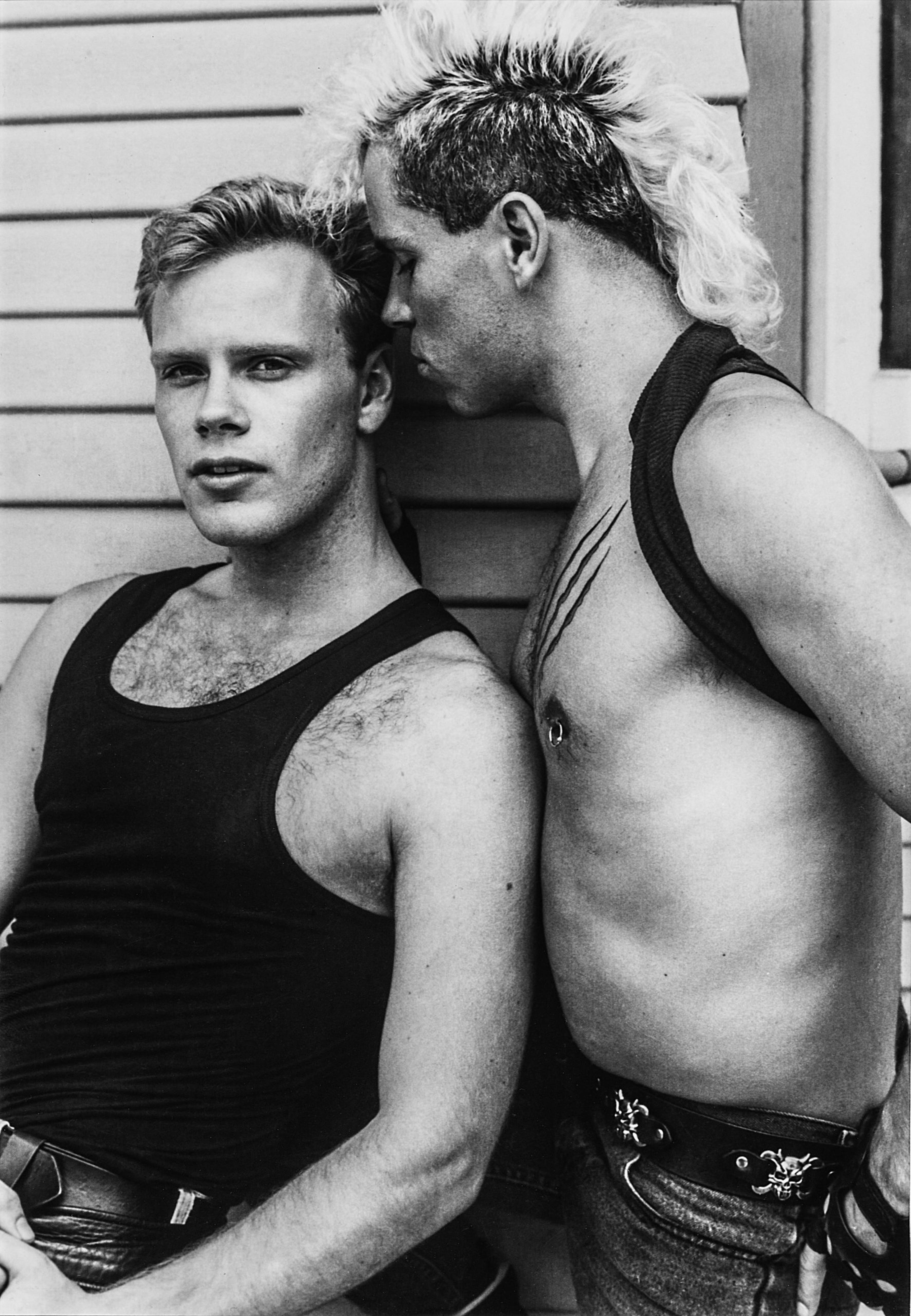 A black and white photograph of a shirtless man with a mohawk whispering to another man in a black tank top