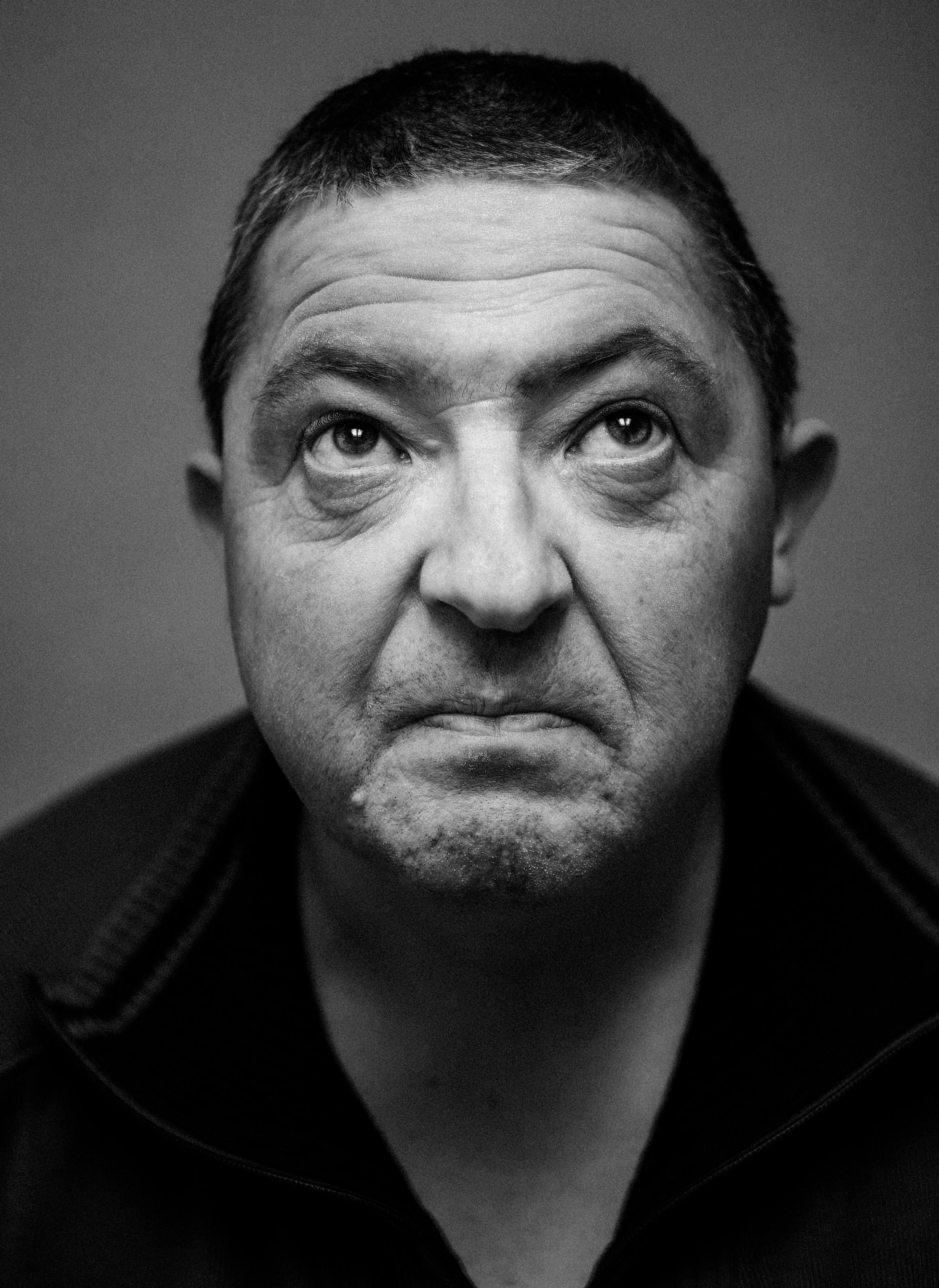 A close-up black-and-white portrait of a middle-aged man with short hair. Captured from a front angle, the image focuses closely on his facial features as he gazes upwards into the light with a thoughtful and solemn expression.