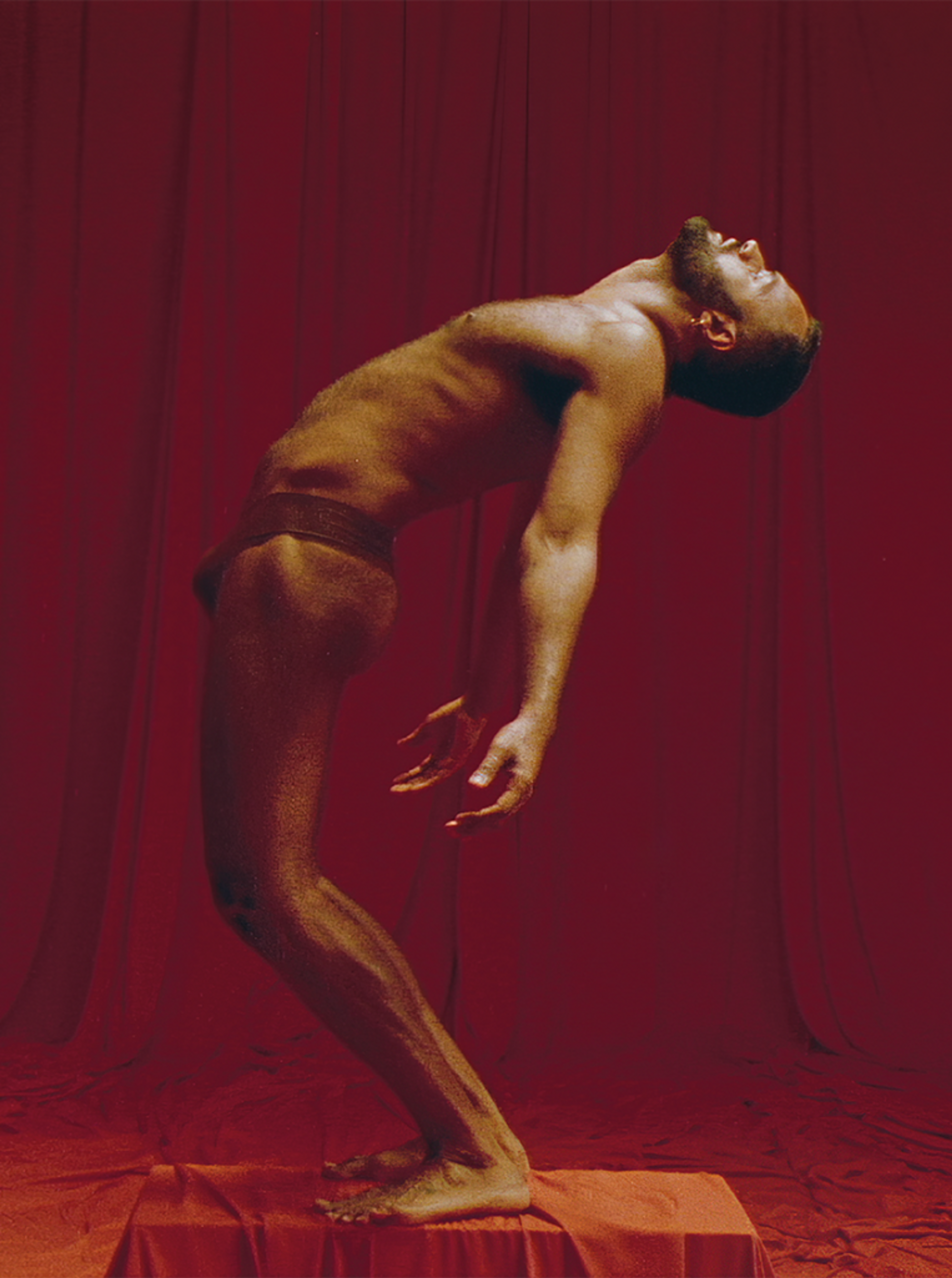 Image of a male identifying figure posing in the nude with a red backdrop.