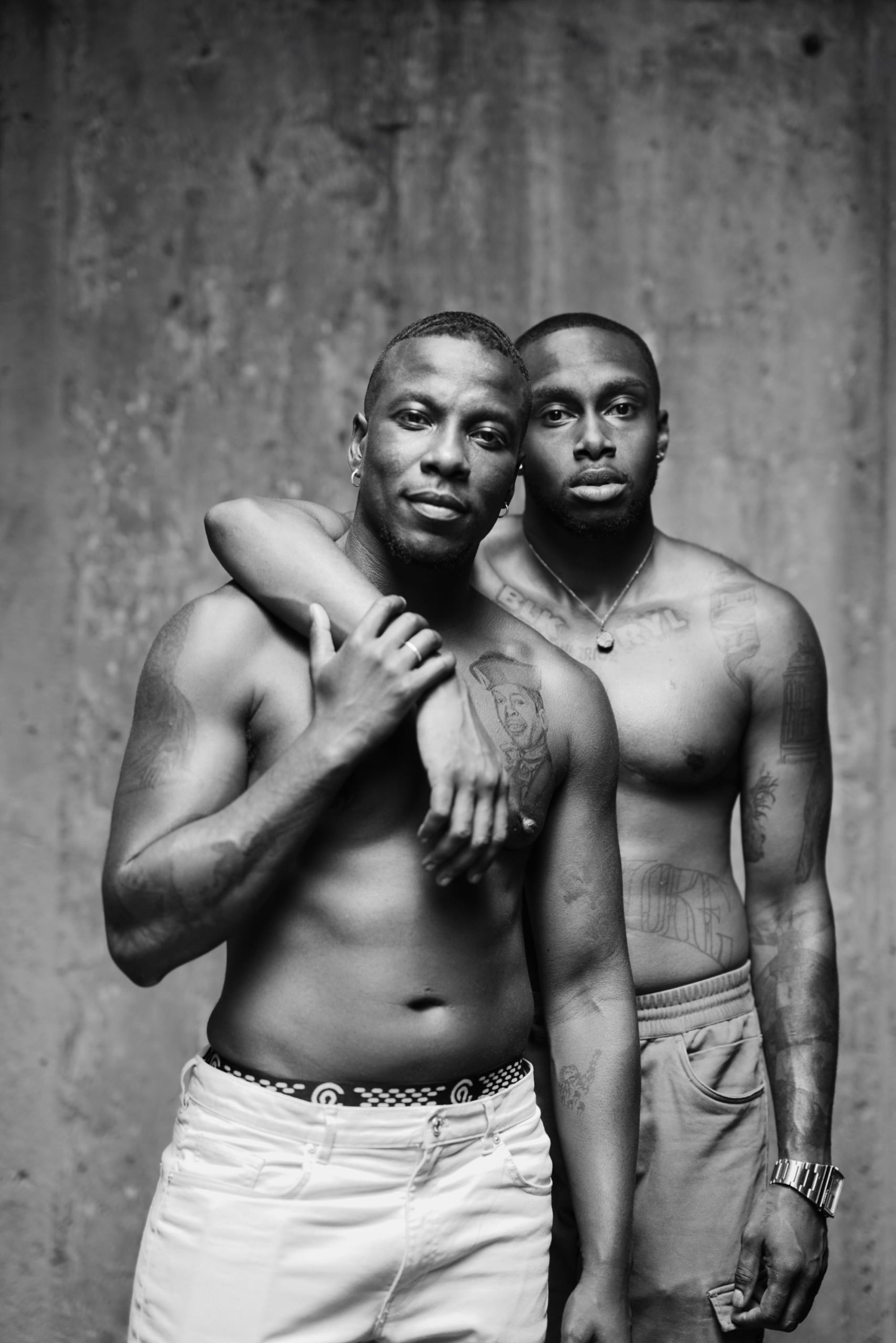 A black and white photograph of two shirtless men, one with his arm draped over the other